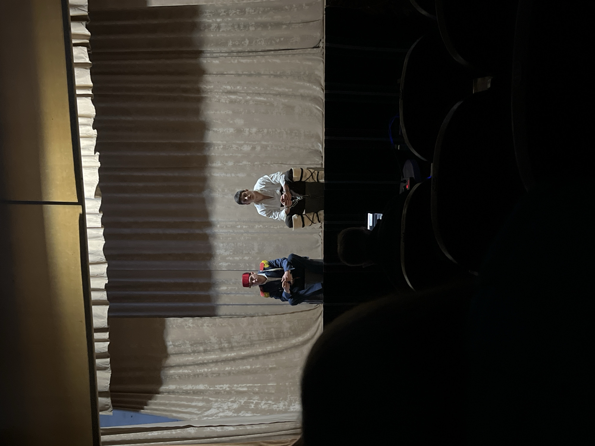 The image captures a theatrical performance viewed from the audience. A beige curtain serves as the backdrop. Two actors are on stage. One actor, wearing a white outfit and leg wrappings, sits on a chair. Chains appear to bind his hands. The other actor stands, wearing a dark blue uniform with gold trim and a red fez. He holds a microphone and seems to be speaking. Seating is visible in the foreground and to the right, suggesting an auditorium setting. The location appears to be a performance in Voiniagovo, Bulgaria.