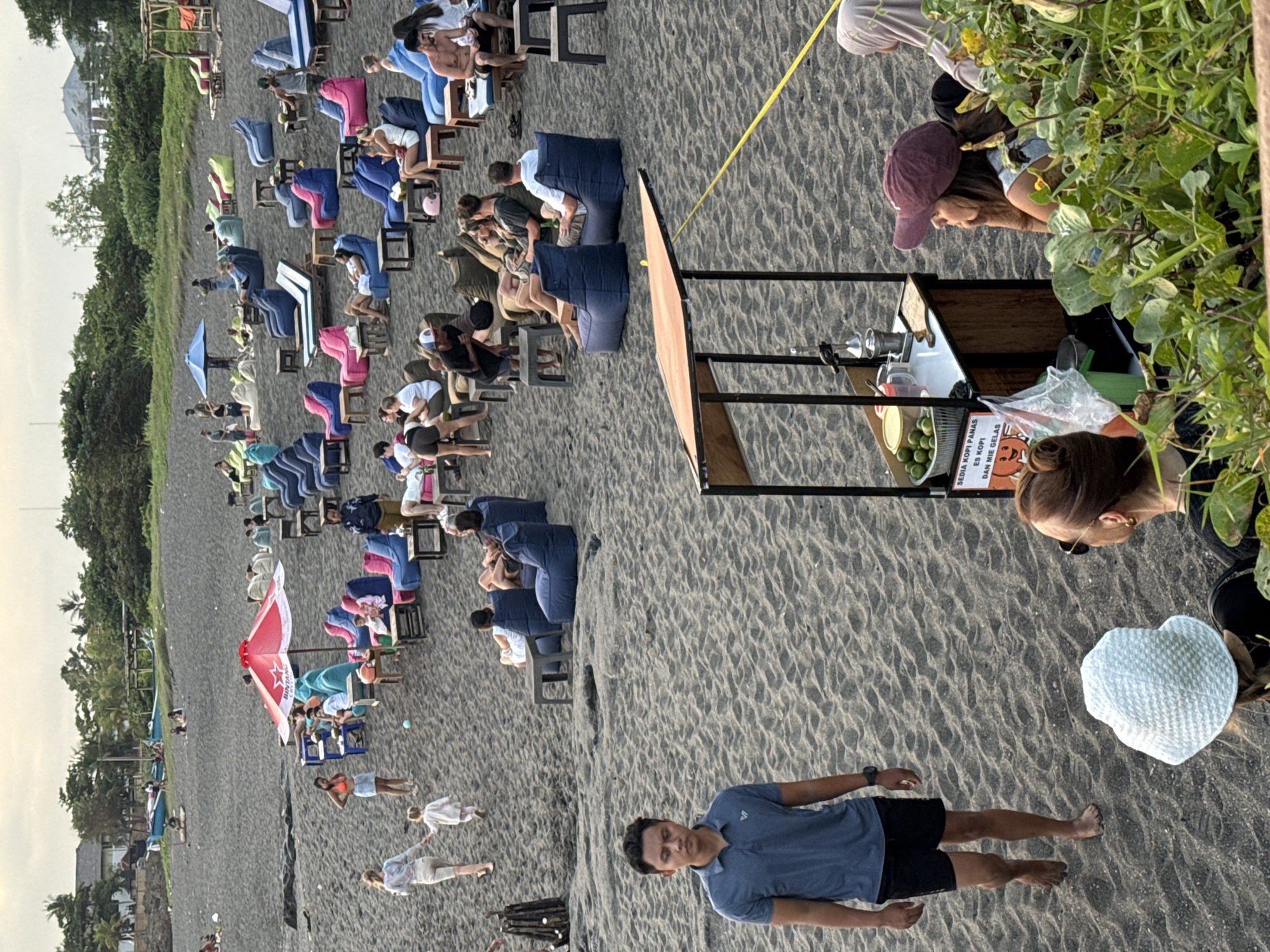 The image depicts a beach in Mengwi, Indonesia. A line of chairs and cushions is arranged on the black sand, presumably for viewing a performance or event. People are seated in the chairs, facing away from the camera. Some people are walking on the beach. A food or beverage stall is situated on the right side of the frame, attended by several individuals. One man stands in the foreground, facing the camera.