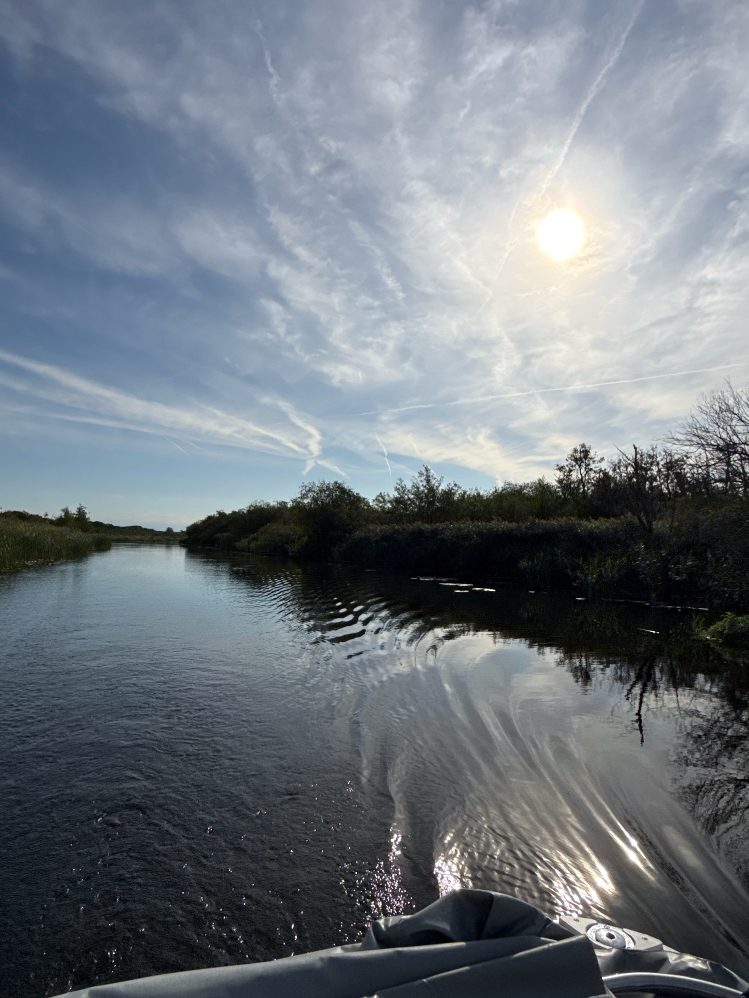 The image depicts a scenic view of a river in North Norfolk, United Kingdom, on a clear day with a bright sun and scattered clouds. The sun is positioned in the upper right quadrant of the sky, casting a strong glare on the water. The river flows from the foreground towards the background, bordered on both sides by lush vegetation including reeds and trees. In the foreground, the bow of a boat is visible, with dark grey fabric covering parts of it. The water exhibits ripples and reflections of the sky and surrounding environment.