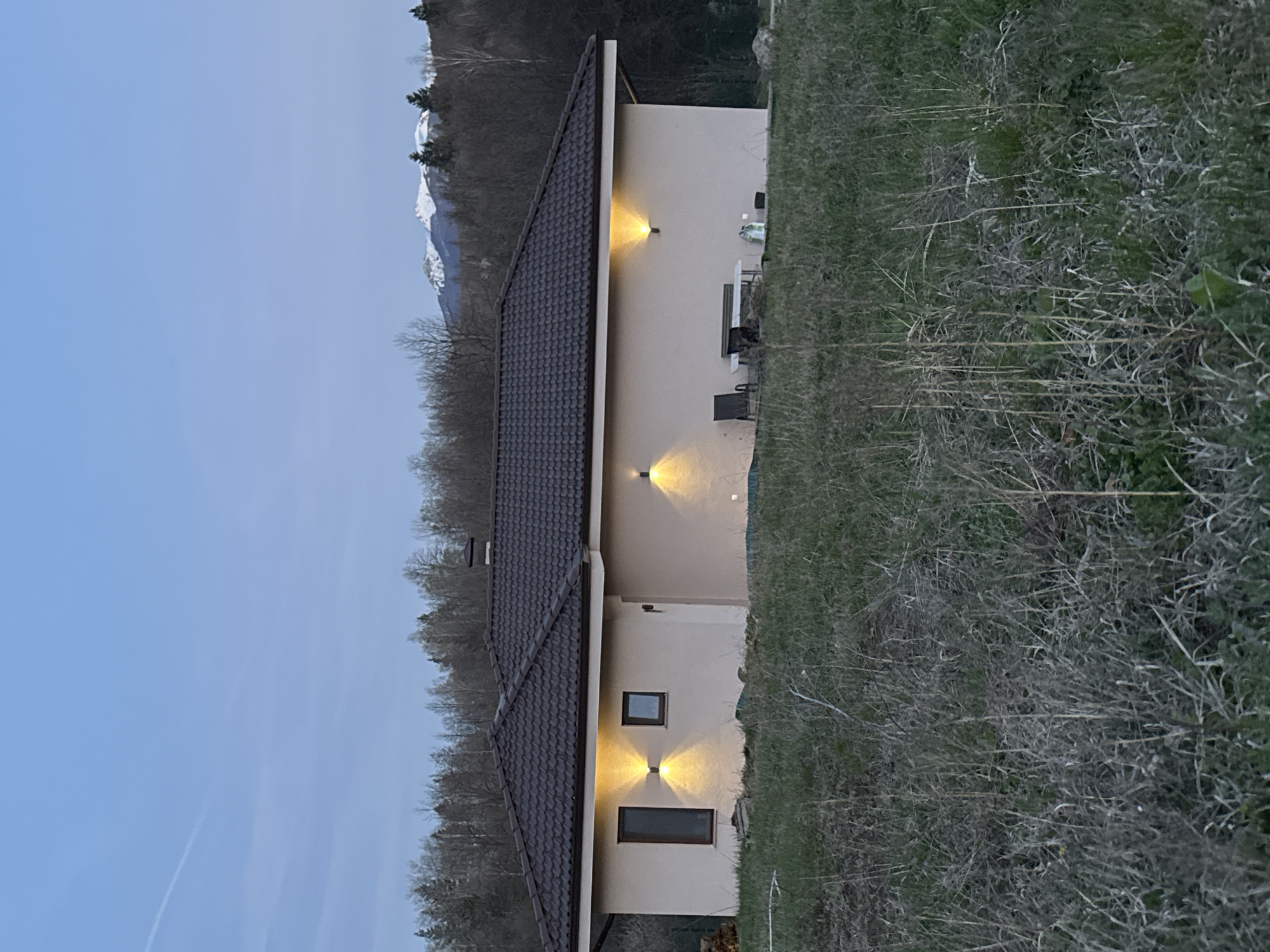 The image depicts a single-story building with a brown tile roof and beige exterior walls. The building is illuminated by three sets of wall-mounted lights. The surrounding environment includes dry, grassy vegetation and a forest with bare trees. In the background, snow-capped mountains are visible. The sky is a pale blue color, suggesting twilight or early evening. No people are visible in the frame. The location is likely in a rural or suburban area near mountains, possibly in the city of Unknown, Unknown.