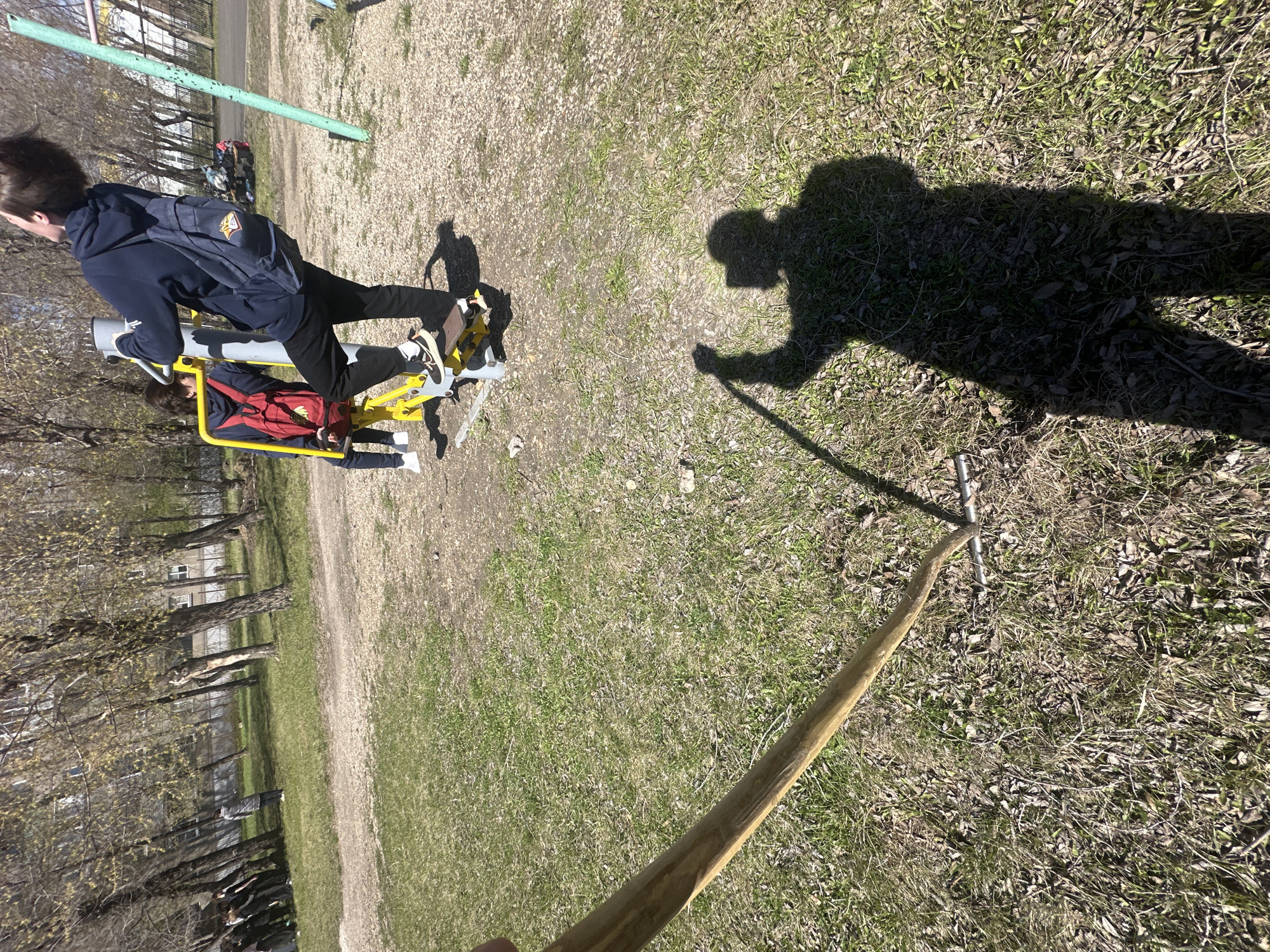An outdoor scene captured on a sunny day depicts a park or schoolyard with sparse grass and dirt paths. The image is taken from a first-person perspective, showing a large shadow of a person holding a long wooden stick with a metal rake head at its end, cast prominently across the grassy ground in the foreground.
In the mid-ground, two individuals are seen from behind, utilizing a yellow and grey outdoor exercise machine, possibly a leg press or similar equipment. One person stands on the machine, wearing a dark blue hoodie, black pants, white sneakers, and a dark blue backpack with a visible yellow patch or logo. The other person is seated on the machine, also wearing dark clothing and a red backpack.
The background features several bare or budding trees, indicating early spring, and a green metal pole that extends vertically. Further in the distance, parts of residential or institutional buildings are visible. The bright sunlight casts long, distinct shadows, suggesting either morning or late afternoon. The ground is a mix of dry grass, patches of dirt, and some scattered dry leaves. No clear legible text is visible on any objects beyond the unreadable logo on the backpack.