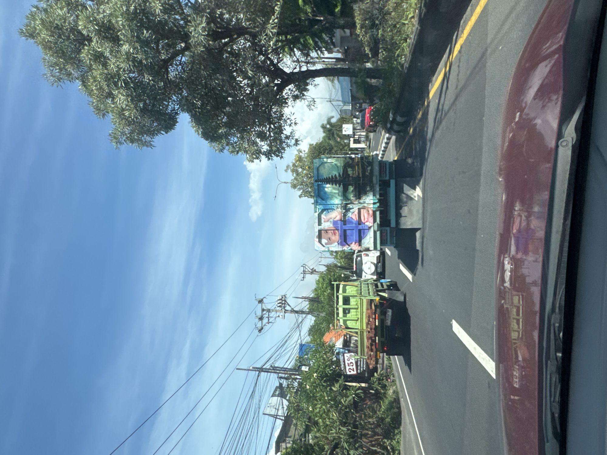 Captured from within a vehicle, the image portrays a street in Denpasar, Indonesia. A dual-lane road with a yellow shoulder stripe is visible, occupied by a line of trucks. The first truck is green with a sign advertising a 25% discount. The subsequent truck is decorated with what appear to be murals. Overhead, utility poles and power lines weave across the sky. Dense trees and foliage line the side of the road, providing a natural backdrop. The vehicle's interior is partially visible on the right side of the frame.