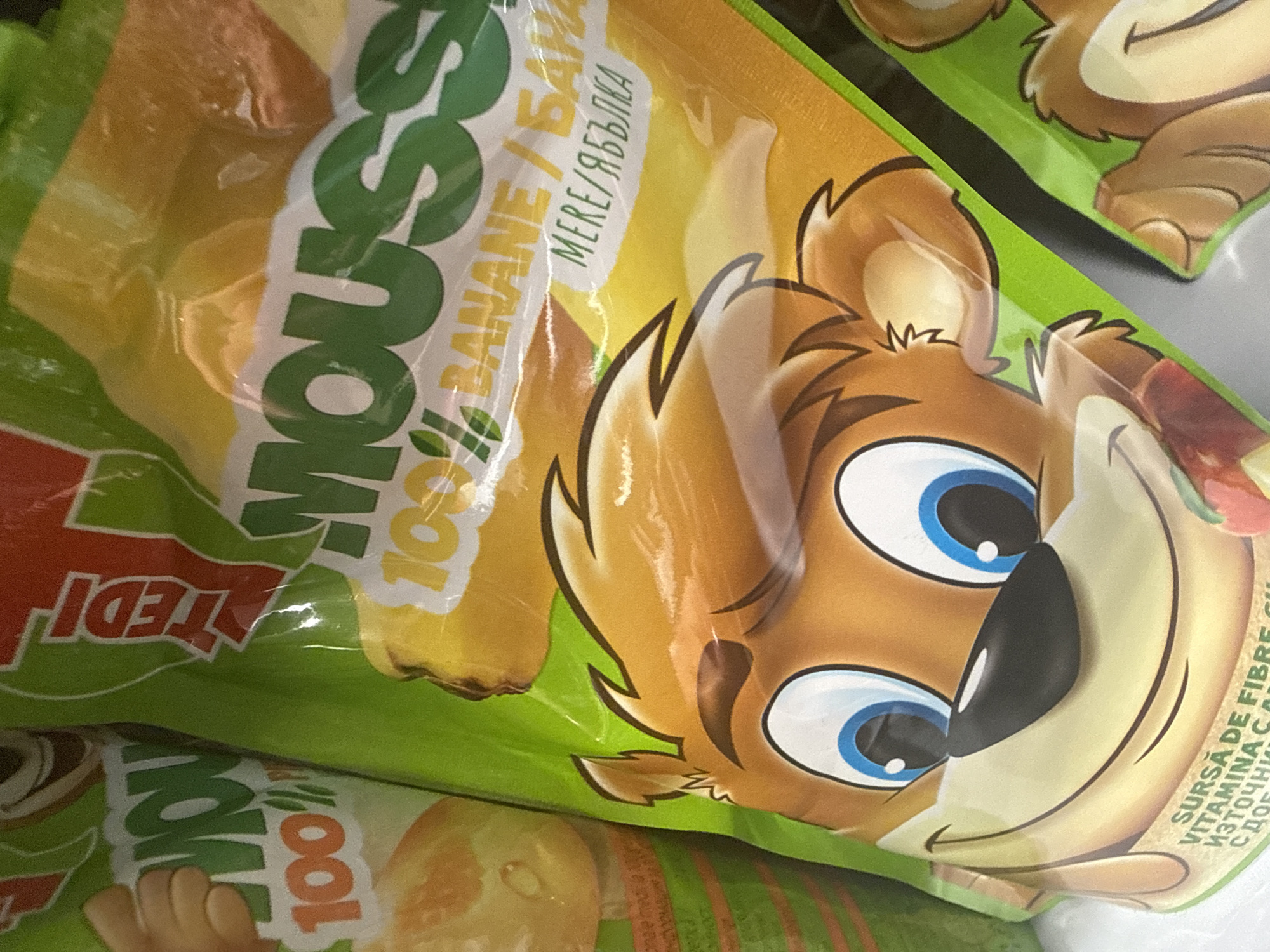 The image shows two bags of "Moussou" brand fruit snacks. The bags are predominantly green and yellow, with cartoon animal faces on the front. The left bag features a cartoon bear-like character with large blue eyes and a smiling mouth, and the text "MOU 100%" is visible on the upper left. The right bag also features a similar cartoon character. The text "MOUSSOU 100% BANANE / БАНАНА" and "MERE / ЯБЪЛКА" is printed on the right bag, indicating banana and apple flavors. The snacks appear to be for children. The image is taken in a retail environment, likely a store shelf in Sofia, Bulgaria, given the Cyrillic text on the packaging and the overall context of consumer goods. There are no people visible in the image. The lighting suggests an indoor setting with artificial light. The packaging is shiny and reflective, indicating it's made of plastic film.