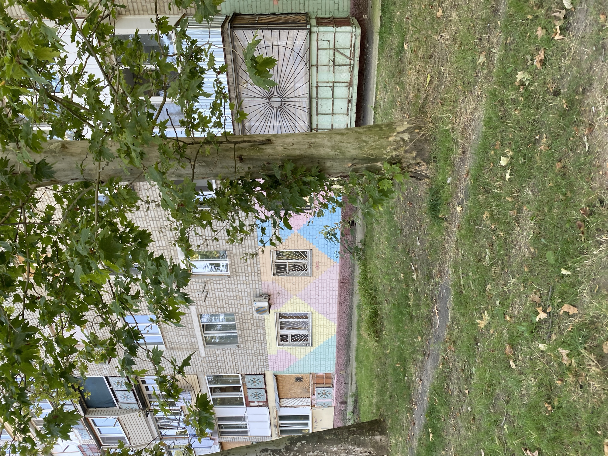 This image captures a section of a brick apartment building in Chornomorsk, Ukraine, partially obscured by the foliage of a large tree. The building's facade is a light-colored brick, with a lower section featuring a colorful diamond-patterned mural in pastel shades of pink, yellow, blue, and green. Several windows of varying sizes are visible, some with metal security bars. An air conditioning unit is mounted on the wall above the mural. To the right, a balcony or enclosed room is visible with a decorative metal grate. The foreground consists of a grassy lawn dotted with fallen leaves, and a dirt path runs through it. The lighting suggests an overcast day.