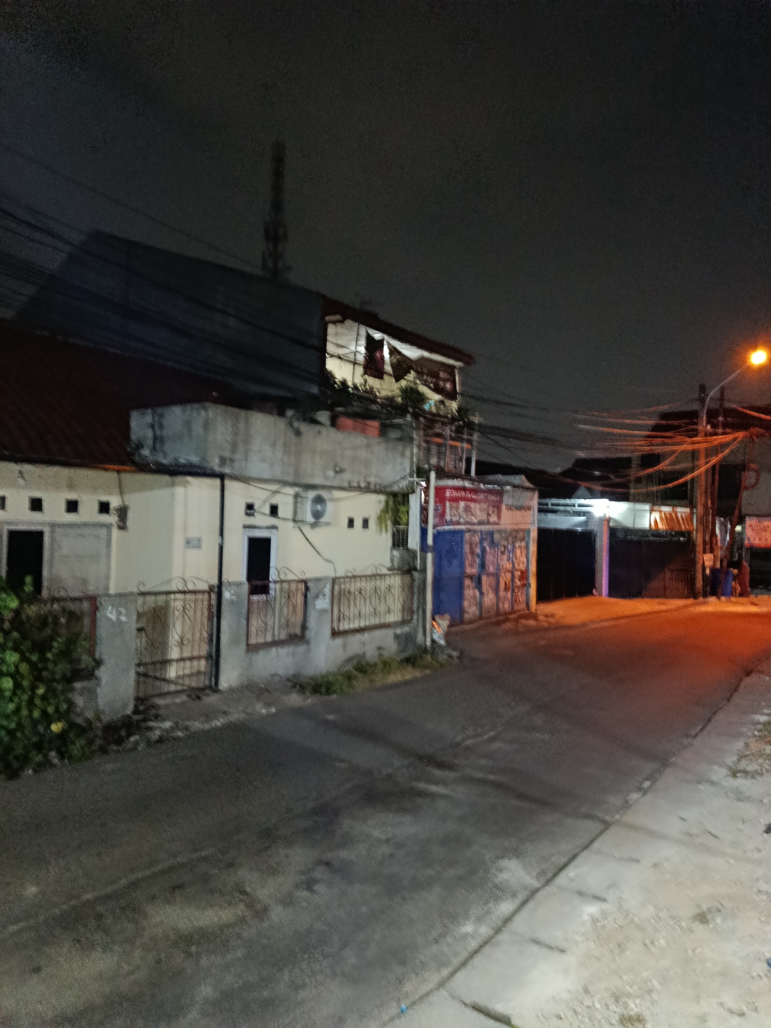 This image depicts a street scene at night in Depok, Indonesia. The setting appears to be a residential or mixed-use area with several buildings visible. In the foreground, a paved street curves to the right, with a concrete sidewalk on the right side of the frame. The street is wet, suggesting recent rain or dew.
On the left side of the street, a single-story building with a red-tiled roof and beige walls is prominent. It has several windows and doors, some of which are covered by metal gates. An air conditioning unit is visible on the exterior wall of this building. Adjacent to the building, there is dense green foliage.
Further down the street, on the right, there are other structures, including what looks like a storefront with blue doors and posters or advertisements on them. Above this, a second-story structure has lights on. A tall telecommunications tower is visible in the distance, silhouetted against the night sky. Overhead, a complex network of electrical wires crisscrosses the scene. A street lamp on the right emits an orange glow, illuminating part of the street.
The time of day is clearly night, as indicated by the darkness and the artificial lighting. The weather is likely damp or has recently been rainy. There are no people actively visible in the frame. The overall mood is quiet and somewhat subdued, characteristic of a nocturnal urban environment.
Visible text includes the number "42" on one of the gates and some text on posters on the blue-doored building. The text on the posters is not clearly legible due to the image quality.