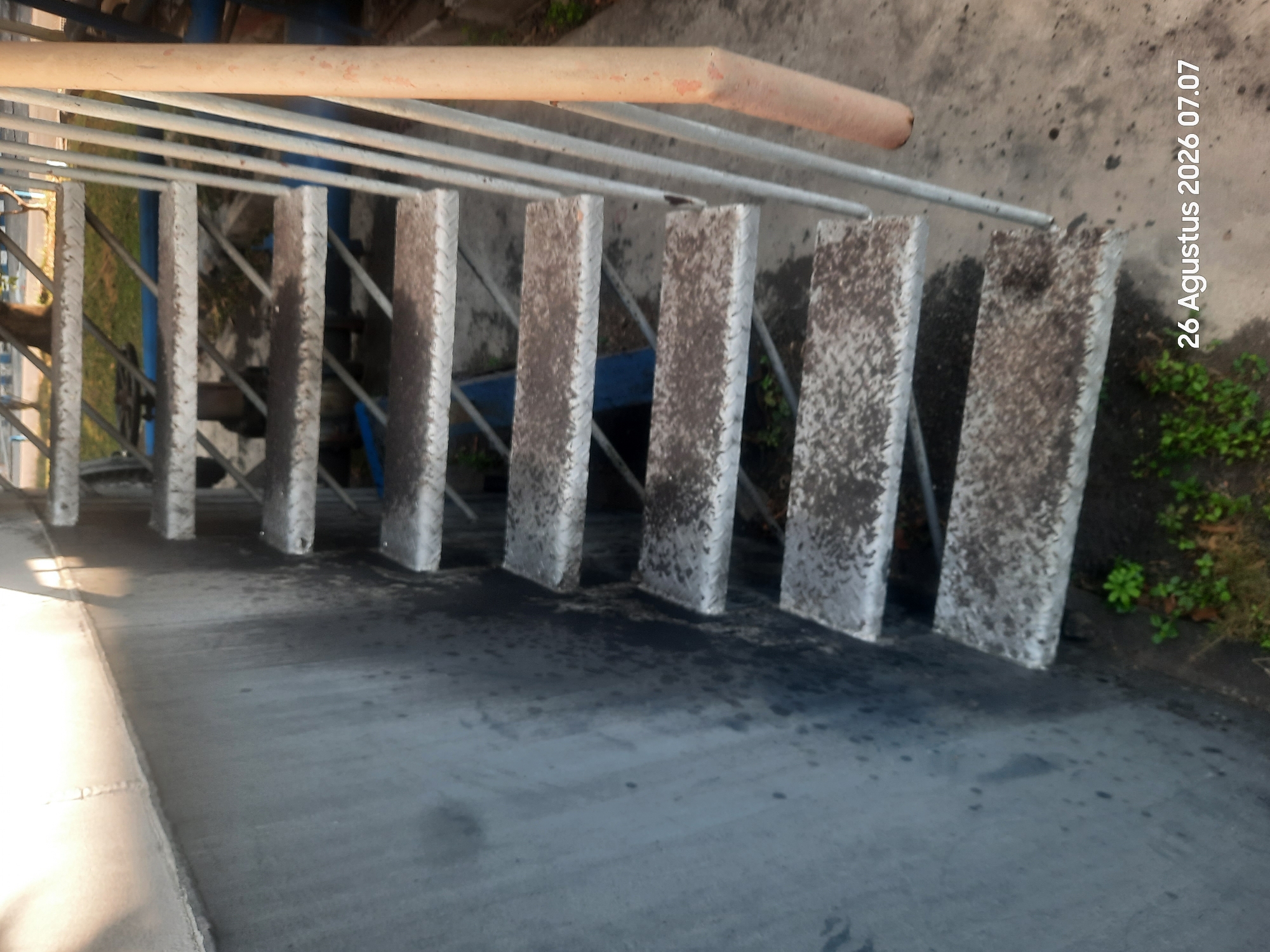 An outdoor, high-angle, close-up shot shows a metal staircase with diamond plate steps. The staircase is attached to a large, cylindrical, metallic structure on the left. The steps are speckled with dirt and debris. A light orange cylindrical object is to the right of the staircase, and metal railings run along the right side of the stairs. The ground below the stairs is concrete with some patches of green vegetation. In the background, out of focus, is some greenery and what appears to be a paved area. The timestamp "26 Agustus 2026 07.07" is visible in the bottom right corner. The scene appears to be in an industrial or utilitarian area in Gending, Indonesia.