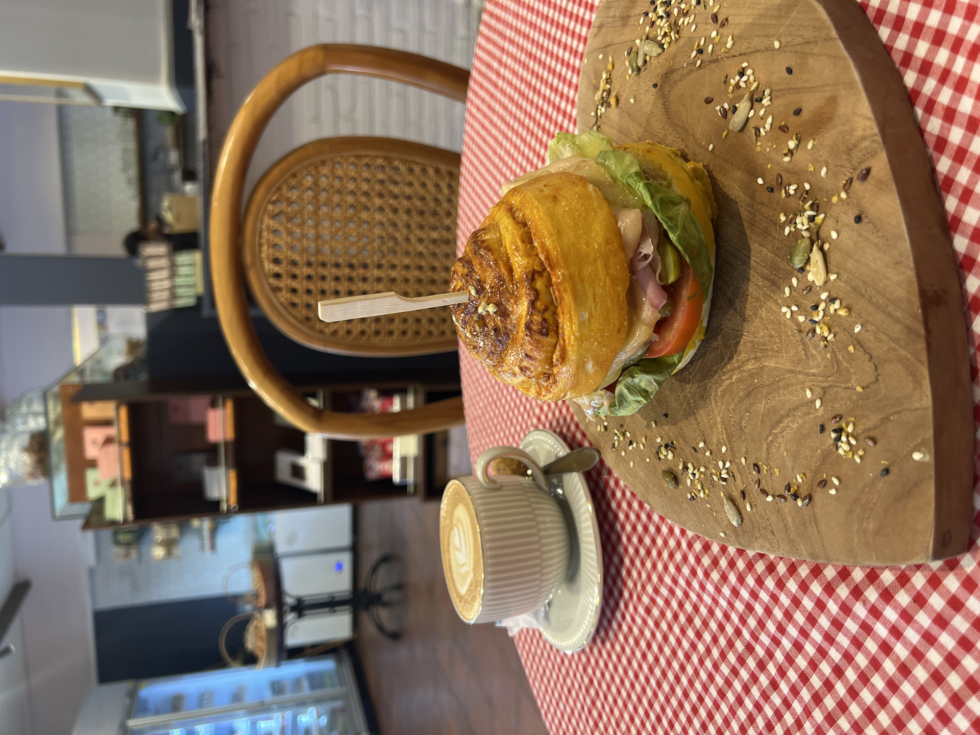 A photograph shows a sandwich and a cup of coffee on a red and white checkered tablecloth. The sandwich is tall and held together with a toothpick. It contains lettuce, tomato, cheese, and meat. It sits on a wooden serving board that is sprinkled with seeds. A cup of coffee with a latte art design is on a saucer next to the serving board. A small spoon rests on the saucer. The scene is set indoors, likely in a cafe or restaurant. A wooden chair is partially visible in the background. Behind the chair, there is shelving with items displayed on it. The photograph was taken in North Kuta, Indonesia. A refrigerated display case is visible near the bottom of the image.