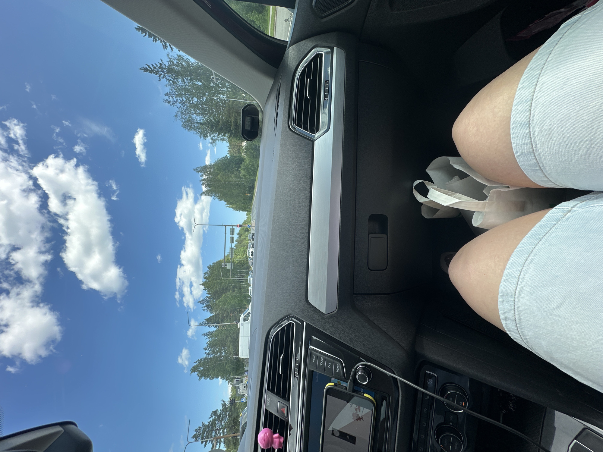 An inside view from a car's passenger seat on a sunny day. The driver's legs, clad in light blue denim shorts, are prominent in the foreground. To the right, a white paper bag rests on the floor. The dashboard is visible, featuring an infotainment screen displaying a navigation app, air vents, and climate control knobs. A pink air freshener is attached to the driver's side air vent. Outside, the car is on a road with trees lining both sides, a blue sky with scattered clouds, and other vehicles in the distance.