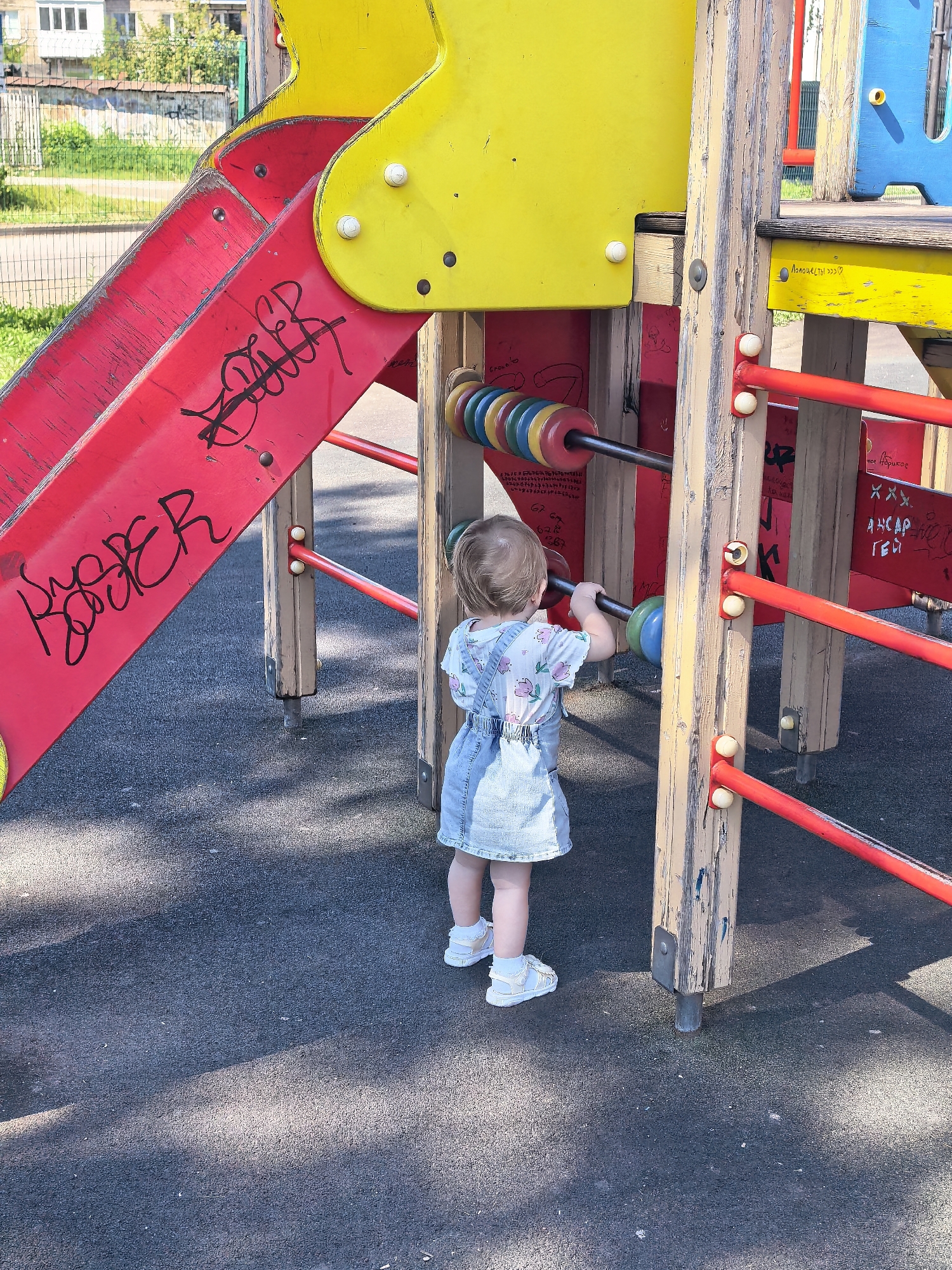 A toddler, viewed from behind, plays with a large, wooden abacus on a bright yellow and red playground structure. The child wears a light-colored dress with a floral pattern and denim overalls, along with white sandals. The abacus features colorful wooden beads threaded onto metal rods. The playground equipment appears to be made of a combination of brightly painted metal and weathered wood. Graffiti is visible on the red slide and on the wooden posts of the structure. The ground is covered in a dark, impact-absorbing material, common in playgrounds. In the background, a metal fence and some greenery are visible, suggesting an outdoor urban setting. The overall lighting indicates a sunny day. The presence of Cyrillic graffiti suggests the location is likely in Russia.