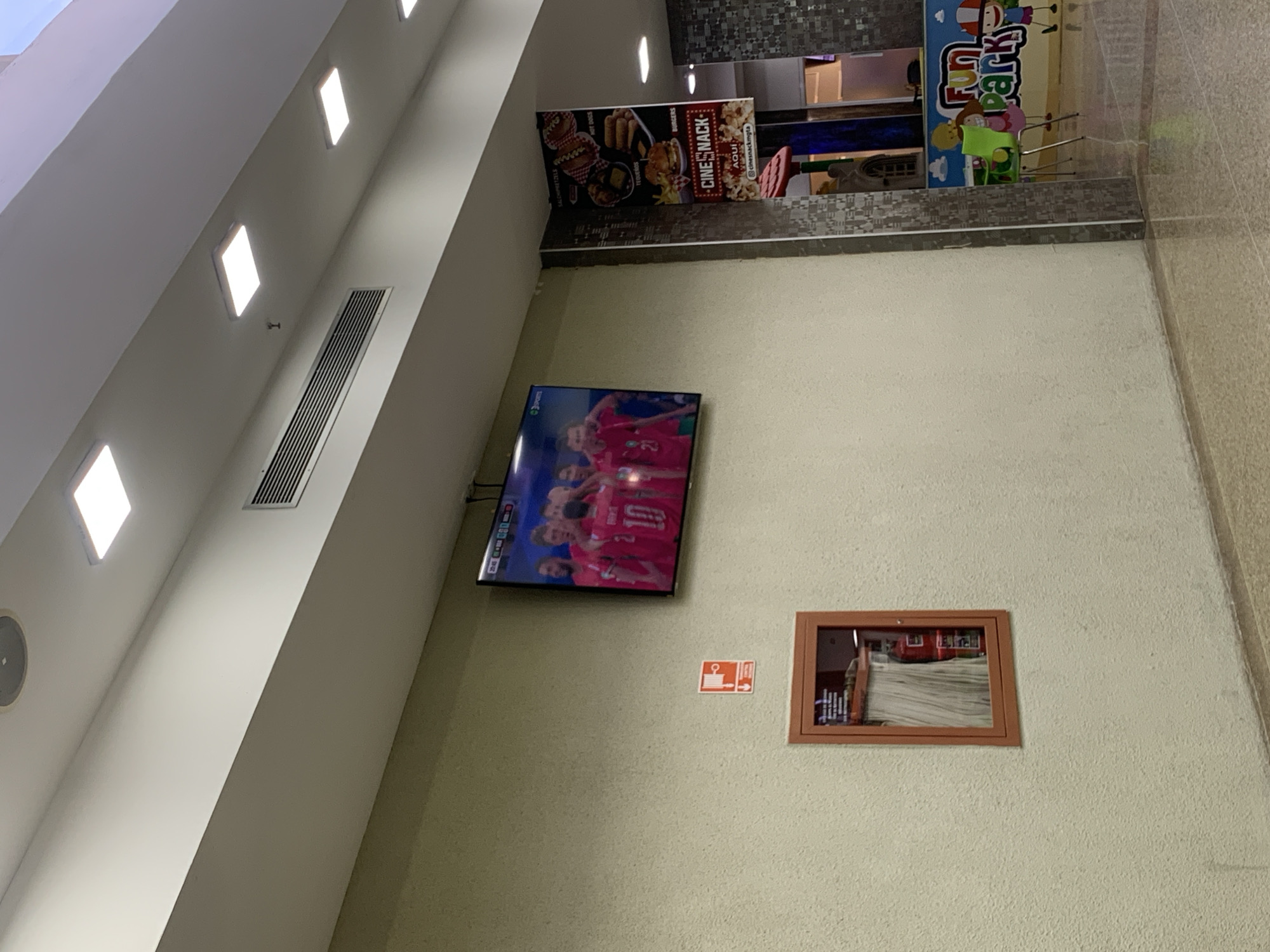 In El Valle del Espíritu Santo, Venezuela, a commercial interior space features a large flat-screen television mounted on a textured, cream-colored wall, displaying a sports event with several players wearing red jerseys. Below the television, a red fire alarm pull station is affixed to the wall. To the left of the alarm, a fire extinguisher cabinet with a glass front contains a red fire extinguisher and coiled hose. Further to the right, a vertical pillar with a gray and black patterned surface separates the wall from a brightly colored "Fun Park" sign. A green plastic chair with chrome legs is visible near the sign. The floor is tiled with a light-colored, speckled pattern, and a slightly darker strip runs along the bottom of the wall. The ceiling, visible at the top, is white with rectangular fluorescent light panels and a ventilation grate. A banner advertising "CineSnack" with images of food items hangs on the pillar.