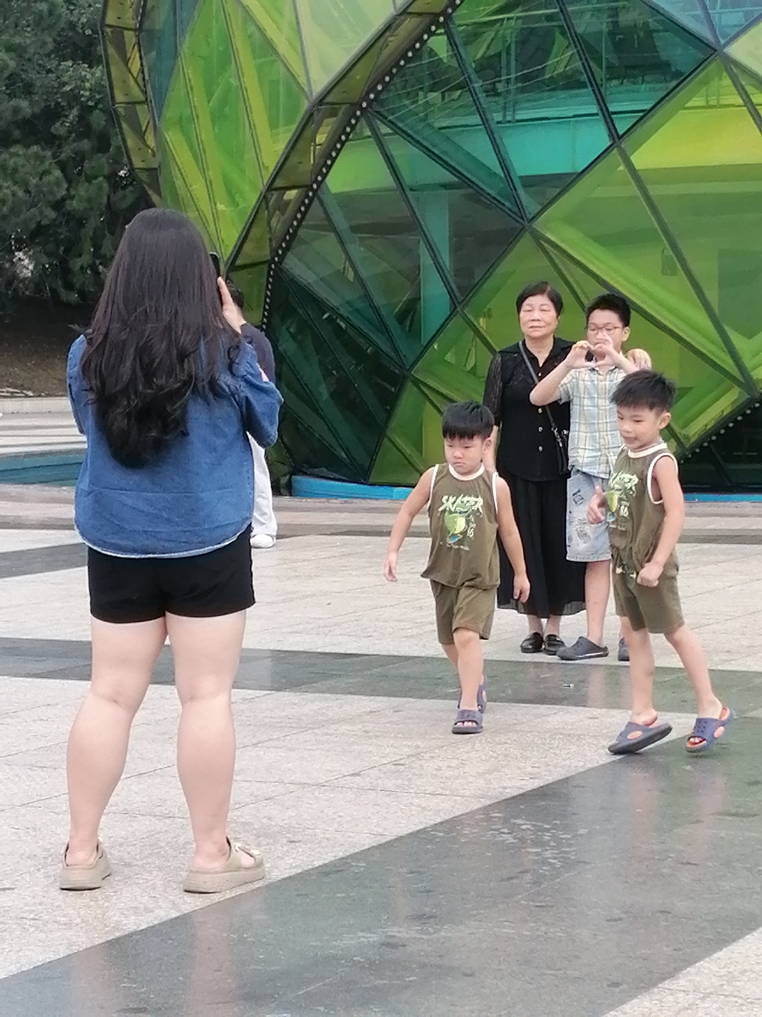 In Da Lat, Vietnam, at the Da Lat Flower Garden, two young boys in olive-green tank tops and shorts are walking. One boy is looking at the camera, while the other is looking forward. Behind them, an Asian woman in a black dress and a young man in a plaid shirt and shorts are posing for a photo. The woman is looking down, and the man is making a heart shape with his hands. In the foreground, a woman with long black hair is taking a photo with her phone. The background features a large, faceted green glass structure, possibly part of an exhibit at the garden. The ground is paved with light-colored tiles, with some darker tiles forming a diagonal pattern.
