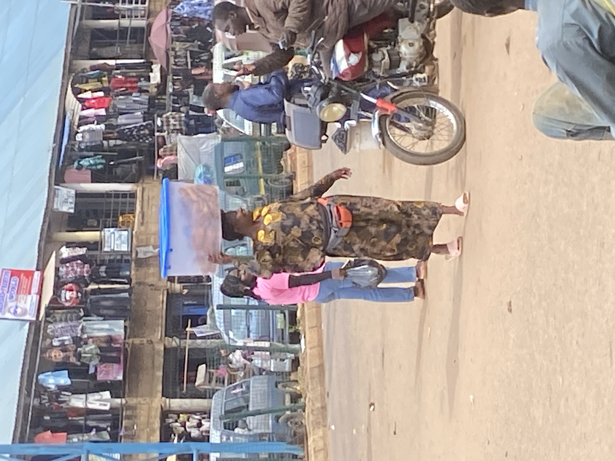 The image depicts a street scene in Bukuru, Nigeria. In the foreground, a woman wearing a patterned dress and blue jeans carries a large blue container on her head. She is walking on a dirt road next to a parked motorcycle. To her left, a stall with shelves filled with clothing is visible. A man in a brown jacket is seen on the motorcycle, and another person in a blue jacket is also near the motorcycle. Further back, more stalls and buildings line the street, with various goods displayed. The sky appears overcast, suggesting a cloudy day. The overall atmosphere seems to be that of a busy marketplace. A red and white sign with text is visible on a building in the background. The time of day is unclear, but the lighting suggests it is daytime.