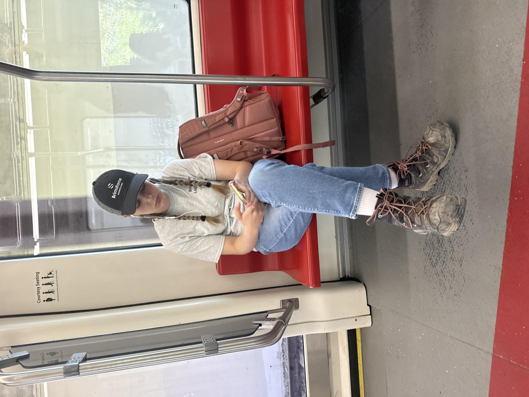 A young person with braided hair and wearing a grey t-shirt, blue jeans, and dirty hiking boots sits on a red transit seat, looking at their phone. They are wearing a black baseball cap with a "Brimstone" logo and white earbuds. A pink backpack is on the seat beside them. A sign indicating "Courtesy Seating" with pictograms of pregnant women, the elderly, and people with disabilities is visible on the wall to the left of the person. The scene takes place on a Calgary Transit train, indicated by the red seats and the overall interior design consistent with Canadian public transit. The floor is grey with scattered debris and a red stripe along the edge. Outside the window, foliage is visible, suggesting an outdoor environment.