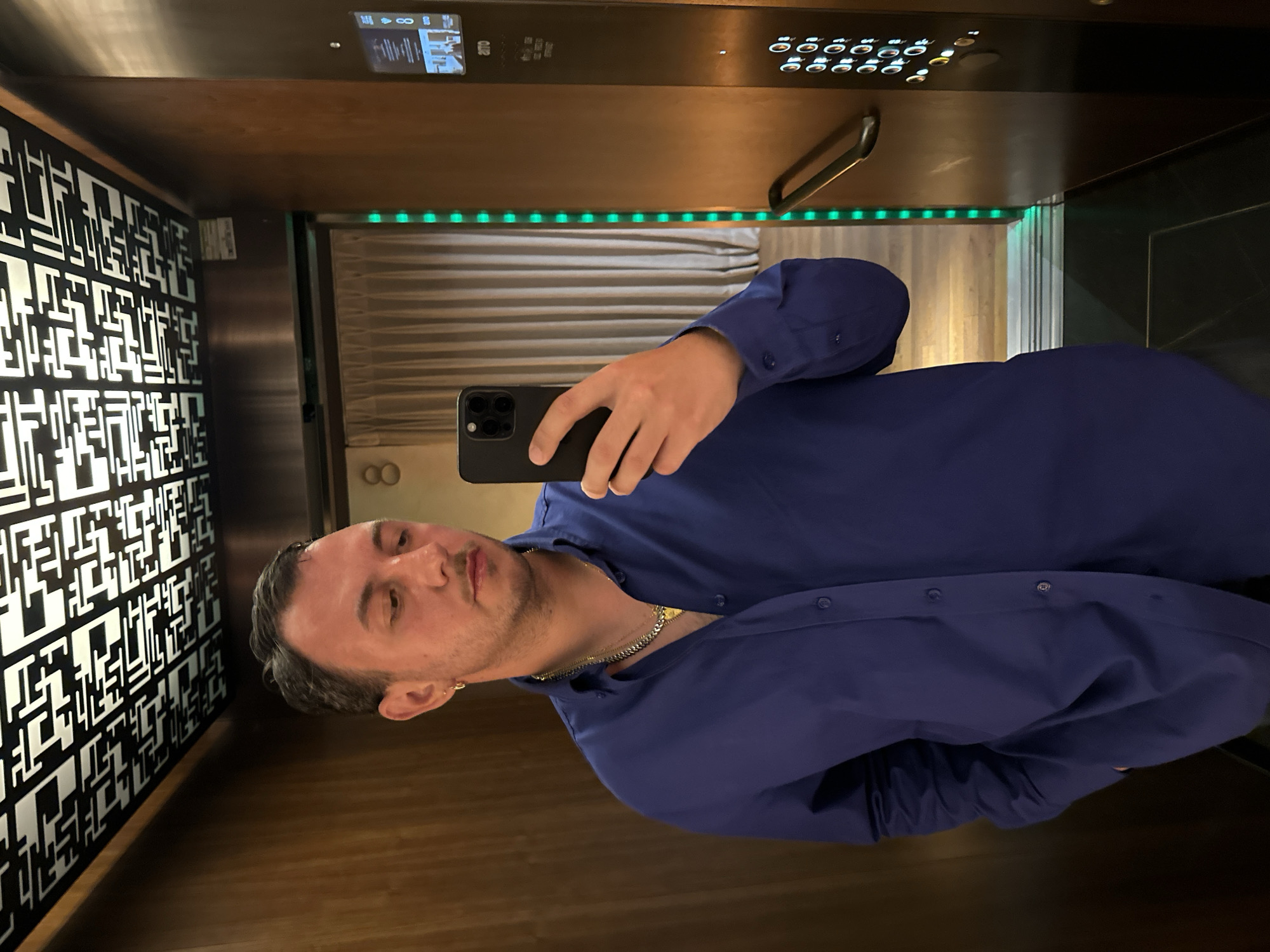 An adult male is captured in a selfie taken inside a modern elevator. He holds a black smartphone in his left hand, pointing it towards his face. The man has dark, slicked-back hair and is wearing a royal blue long-sleeved button-up shirt. A gold chain necklace with a small pendant is visible around his neck, and he has an earring in his left ear. He looks directly at the camera with a neutral expression and slightly pursed lips.
The elevator interior is stylish, featuring distinct design elements. The wall to the man's left is adorned with a striking black-and-white geometric pattern, composed of interconnected lines and shapes. The floor appears to be made of wood or a wood-patterned material. Across the back wall, a series of horizontal blinds or textured panels are visible, topped by a strip of bright green LED lights. The right wall and ceiling are highly reflective, creating mirrored distortions of the man and the opposing patterned wall.
A contemporary elevator control panel is situated on the right side, featuring a row of circular white buttons, some of which are illuminated. A digital display on the panel shows the number "88" along with several small icons, including what appears to be a Wi-Fi symbol. Some small, unreadable text and the letters "ATN" are also discernible on the panel. A metallic handrail is mounted on the reflective right wall. The setting suggests an upscale environment, consistent with a modern building in a city like Madrid, Spain.
