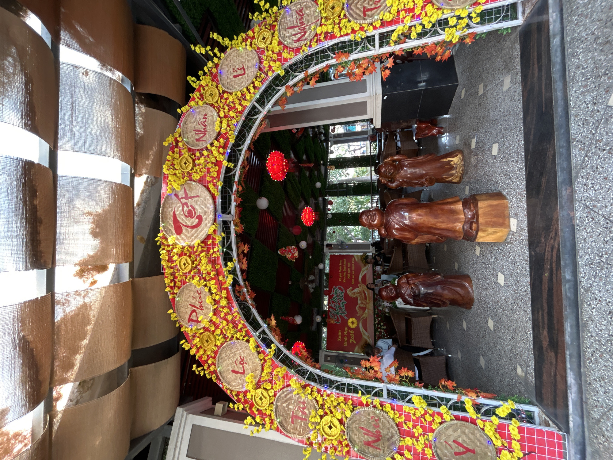 The image shows an indoor archway decorated with Vietnamese New Year (Tet) elements. The arch is white with yellow flowers, straw hats, and red accents, typical for Tet decorations. The words written on the hats are likely greetings related to the New Year.
Inside the archway, there are wooden statues of figures that may represent monks or sages, commonly found in Vietnamese decor. Beyond the statues is the interior of what appears to be a cafe or restaurant, with tables, chairs, greenery, and red lanterns. A sign with Vietnamese text can be seen, possibly related to the cafe's name or Tet wishes.
Given the decor and sign's language, the location is likely in Vietnam, potentially in a tourist area preparing for Tet. Nha Trang, Vietnam, is known to have a "Vườn Treo" cafe, which means "Hanging Garden" in Vietnamese, which is reflected on the sign.