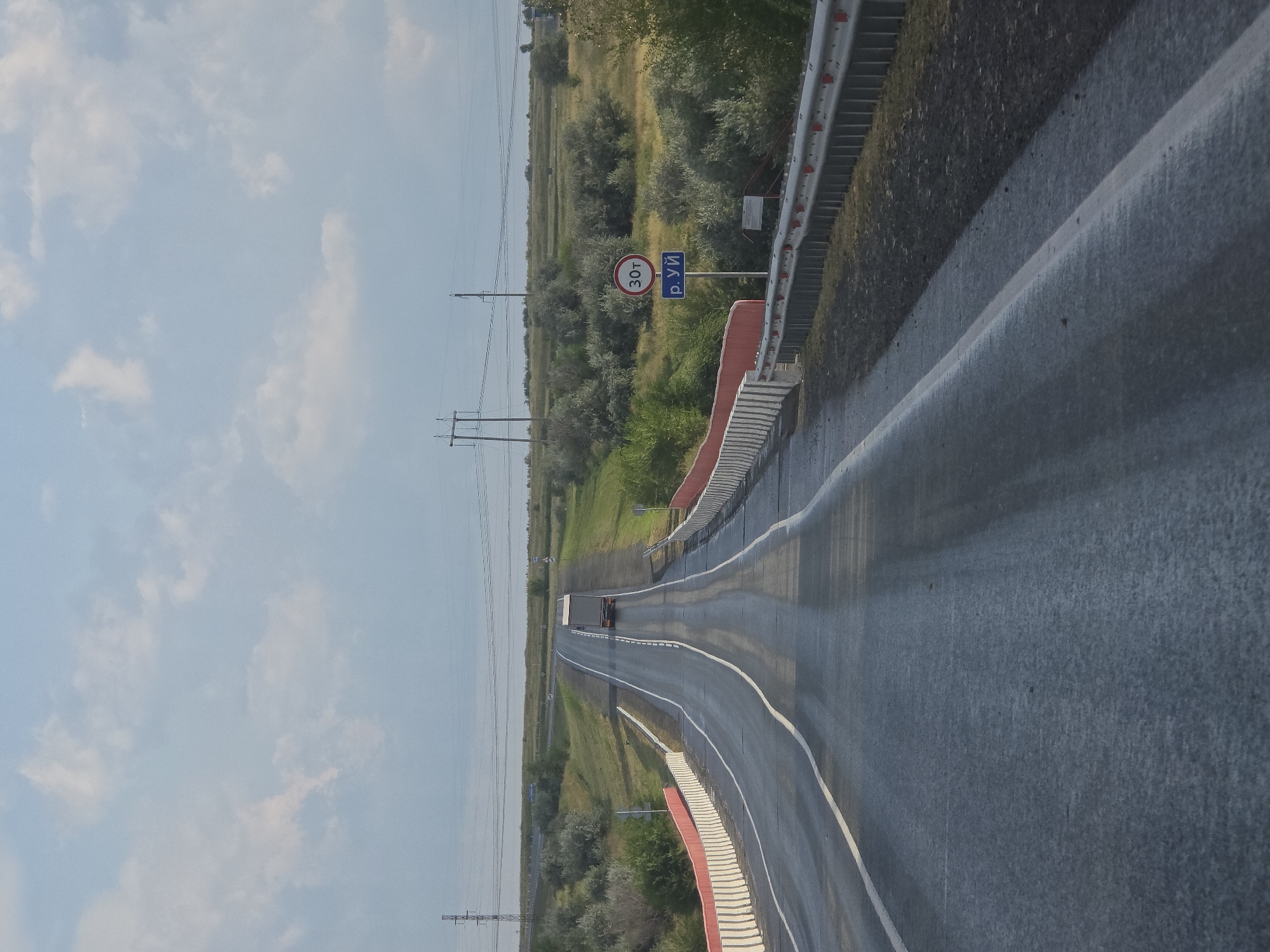 A view from a vehicle driving on a wet asphalt road heading uphill towards a truck. The road has white lane markings and is bordered by a red and white barrier on the left and a silver metal guardrail on the right. Beyond the road, the landscape is covered in green and yellow vegetation, with scattered trees and shrubs. In the distance, power lines and poles are visible against a cloudy sky. A circular traffic sign with "30т" in red and a blue sign with "р. УЙ" are mounted on a pole to the right of the road. The scene appears to be in the Troitsk urban district, Russia, suggesting a rural or semi-rural highway.