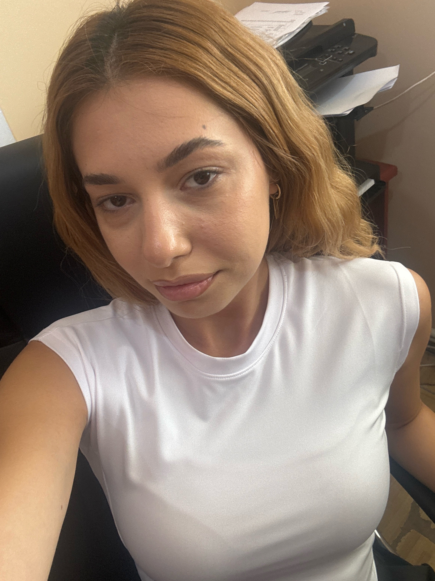 A young woman with shoulder-length auburn hair and light brown eyes is looking at the camera. She is wearing a white t-shirt. Her face is centered in the frame, with her head tilted slightly to her left. Her skin appears smooth and lightly tanned. She has on subtle makeup, with defined eyebrows and a neutral-colored lipstick. She is wearing a small gold hoop earring in her visible ear.
In the background, on the right side of the image, a black office printer is visible, with stacks of paper on top of it. The wall behind the printer is a light peach color. The woman is seated in a dark office chair, which takes up the left side of the frame.
The image appears to be taken indoors, likely in an office environment. The lighting is natural and soft, creating subtle shadows on the woman's face. There is no specific information about the location being Ijevan, Armenia, as this detail is not discernible from the visual content of the image alone.