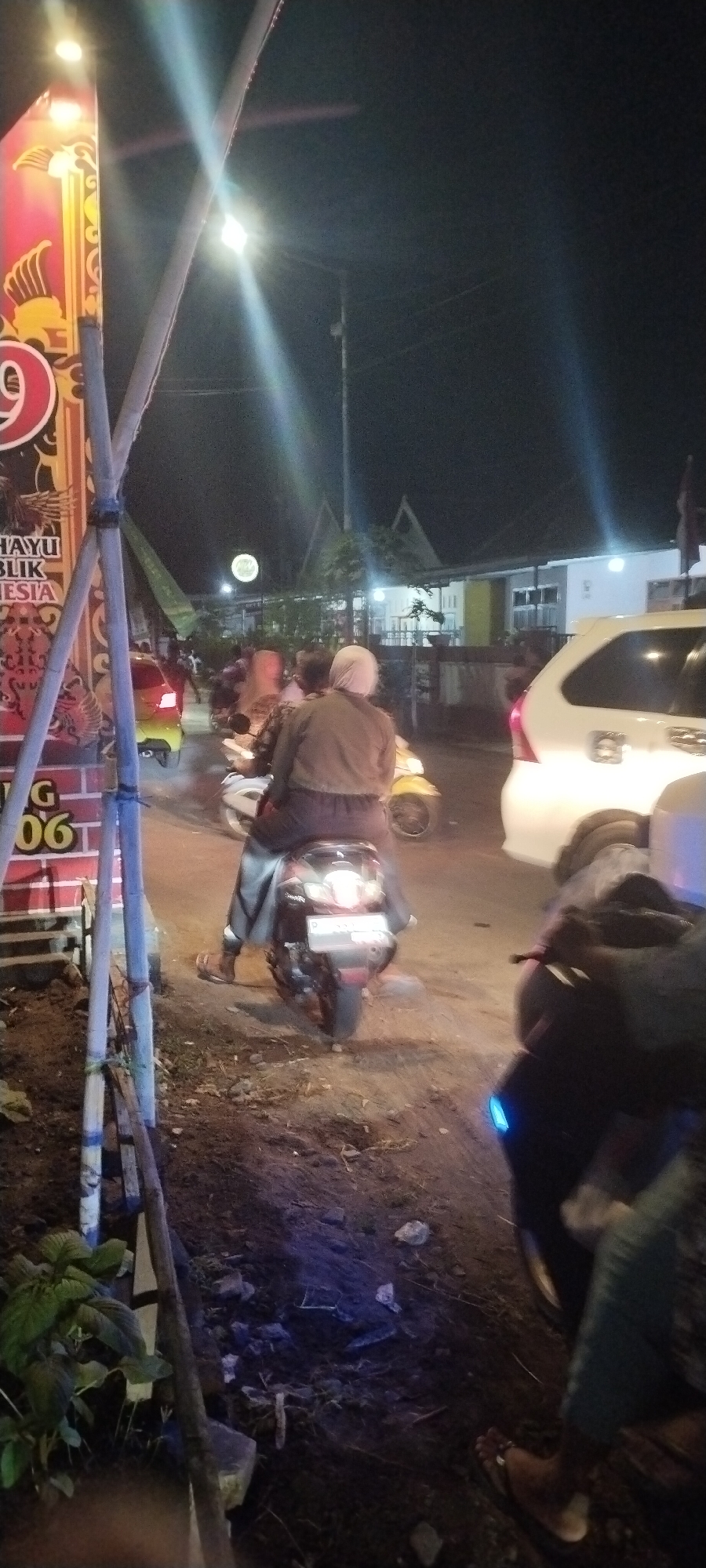The image depicts a nighttime street scene in Keting, Indonesia, likely a residential or commercial area given the presence of houses and a store. In the foreground, a dark-colored motorcycle is parked, its blue lights visible. To the left, a vertical sign with red and yellow decorative elements, possibly advertising, stands prominently. Part of the sign shows text like "HAYU BLIK ESIA" and "G 06". A tall utility pole with wires crosses the scene.
In the middle ground, two individuals are riding a motorcycle. The person in the rear is wearing a light brown jacket and a white head covering, while the person in front has a darker jacket and is obscured. The motorcycle they are riding is dark and has a license plate. To their left, a yellow car is visible.
Further back, several other motorcycles and at least one white car are present, suggesting moderate traffic. The buildings in the background appear to be low-rise structures with illuminated windows and distinctive pitched roofs. The overall environment is dimly lit, with streetlights and vehicle lights providing illumination. The ground appears to be unpaved in some areas, with dirt and scattered rocks. A small green plant is visible in the lower left corner. The atmosphere is typical of a busy street at night in a populated area.