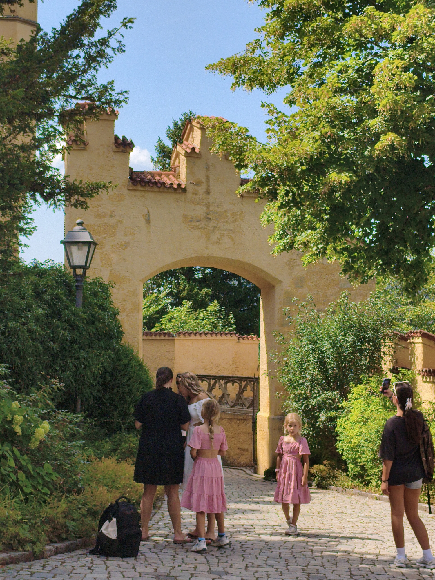 The image shows a paved pathway leading to a yellow arched gateway of a castle or historic building, possibly Hohenschwangau Castle, in Schwangau, Germany. Lush green trees and foliage flank the pathway and surround the gateway.
In the foreground, a family group is visible. A woman in a black dress stands to the left, facing away from the camera, with a backpack at her feet. Beside her are two young girls in pink dresses. The girl on the right is slightly ahead and facing the camera. To the far right, another individual in a dark top and shorts is taking a photo with a smartphone. The pathway is made of cobblestones. The sky above is a clear blue with a few wispy clouds. The architecture of the gateway features decorative crenellations and an arched opening that reveals more greenery behind it. A vintage-style lamppost stands to the left of the pathway, near the dense foliage.