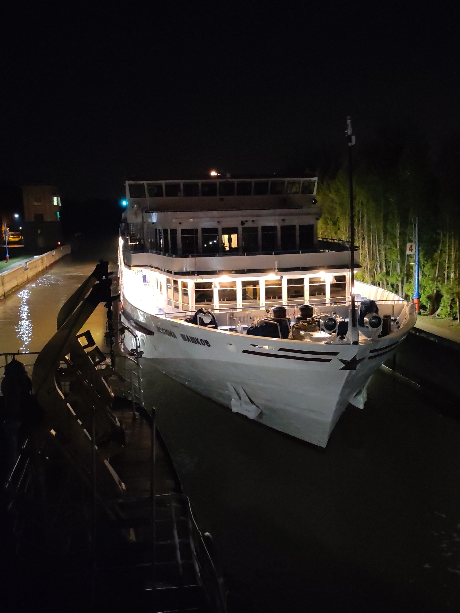 A photograph taken at night depicts the "Rostislav Shashkov" river cruise ship situated within a lock on the Moscow Canal in Dedenevo, Russia. The ship's illuminated multi-tiered structure is seen against the dark background. Locks are visible on both sides of the ship. Trees are behind the lock on the right side. A building is partially visible in the far background on the left. The photograph is shot from another boat on the left side of the river cruise ship.
