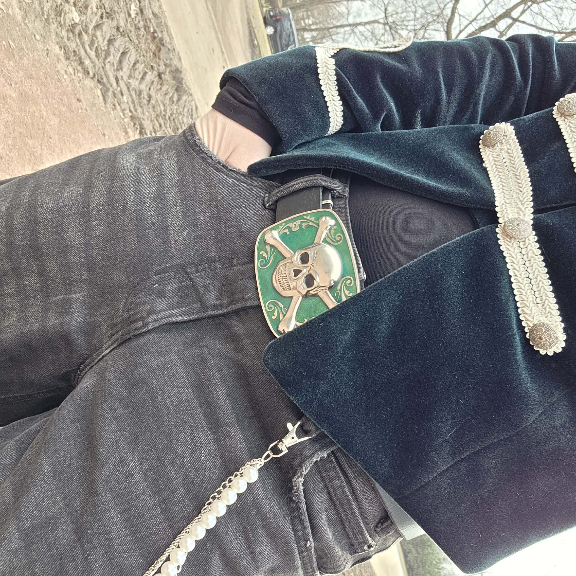 A person is wearing a dark green velvet jacket with white trim and decorative buttons, black pants, and a black belt with a green and gold skull buckle. A pearl chain hangs from the pants. The location appears to be outdoors, with a dirt path visible and trees in the background. The image does not contain enough information to conclusively confirm the location as Saint Petersburg, Russia.