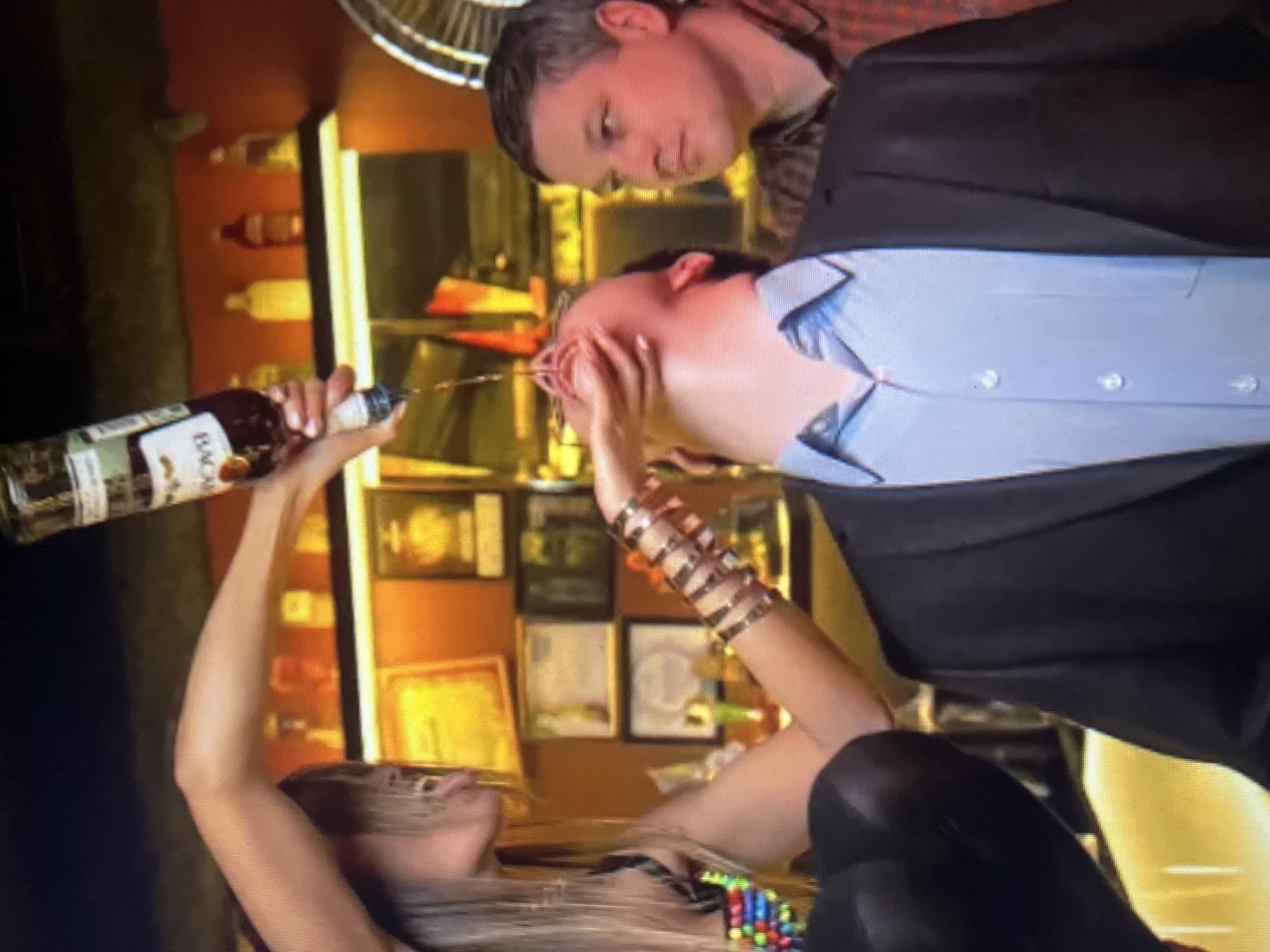 A man in a suit and blue shirt is being poured liquid from a Bacardi bottle by a woman with blonde hair and colorful clothing. The man's head is tilted back and his mouth is open to receive the liquid. Another man in a plaid shirt is looking on from the right. The background appears to be a bar area with shelves of bottles and framed items on the wall. The location is the city administration building of Qarağandı, Kazakhstan.
