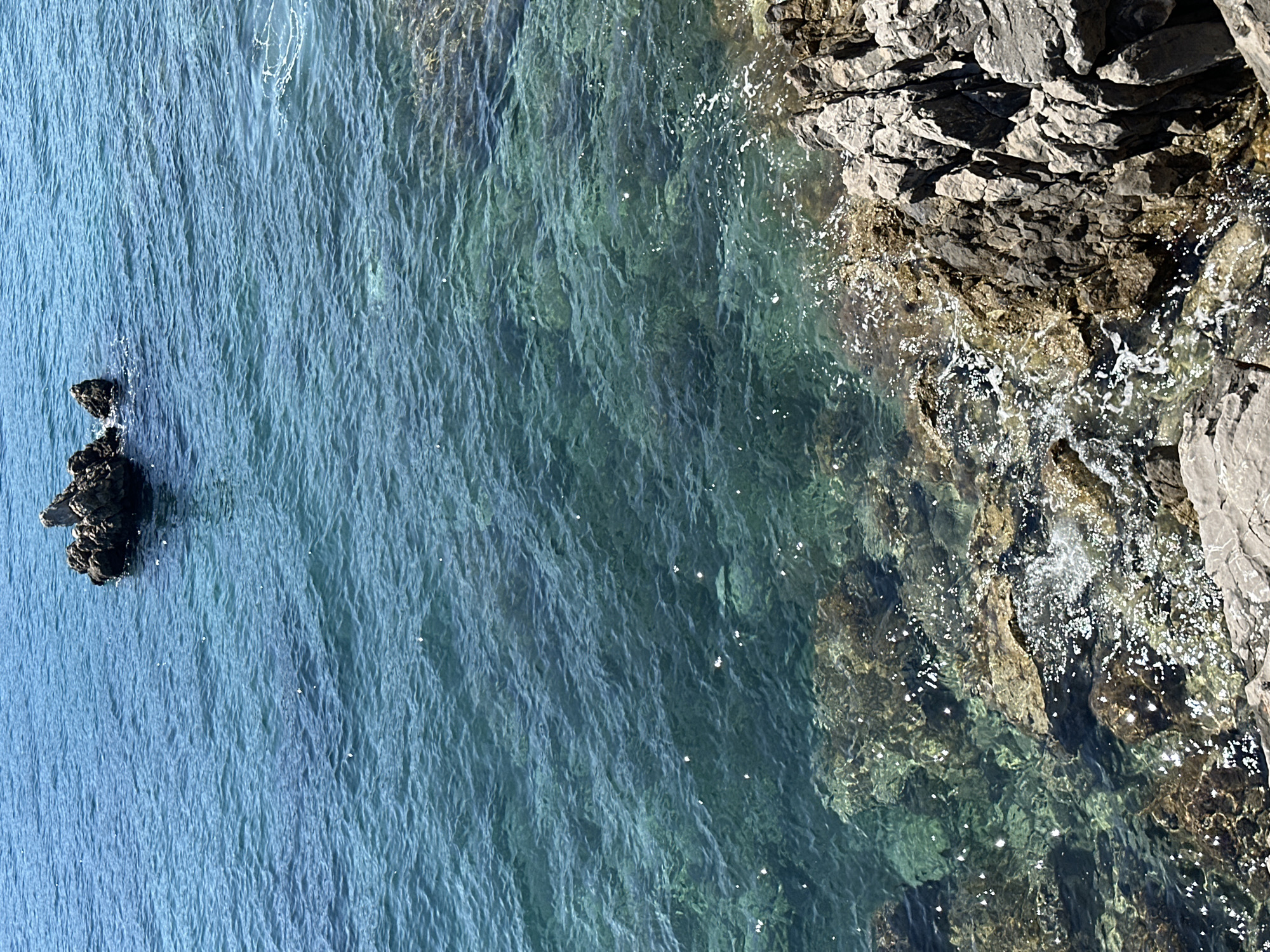 The image depicts a rocky coastline along clear turquoise waters. Rocks line the right edge of the frame, with some extending into the water. An isolated cluster of dark rocks is visible in the upper left of the image. This photograph could be taken on a coastline in Budva, Montenegro, due to its rocky shores and clear water.