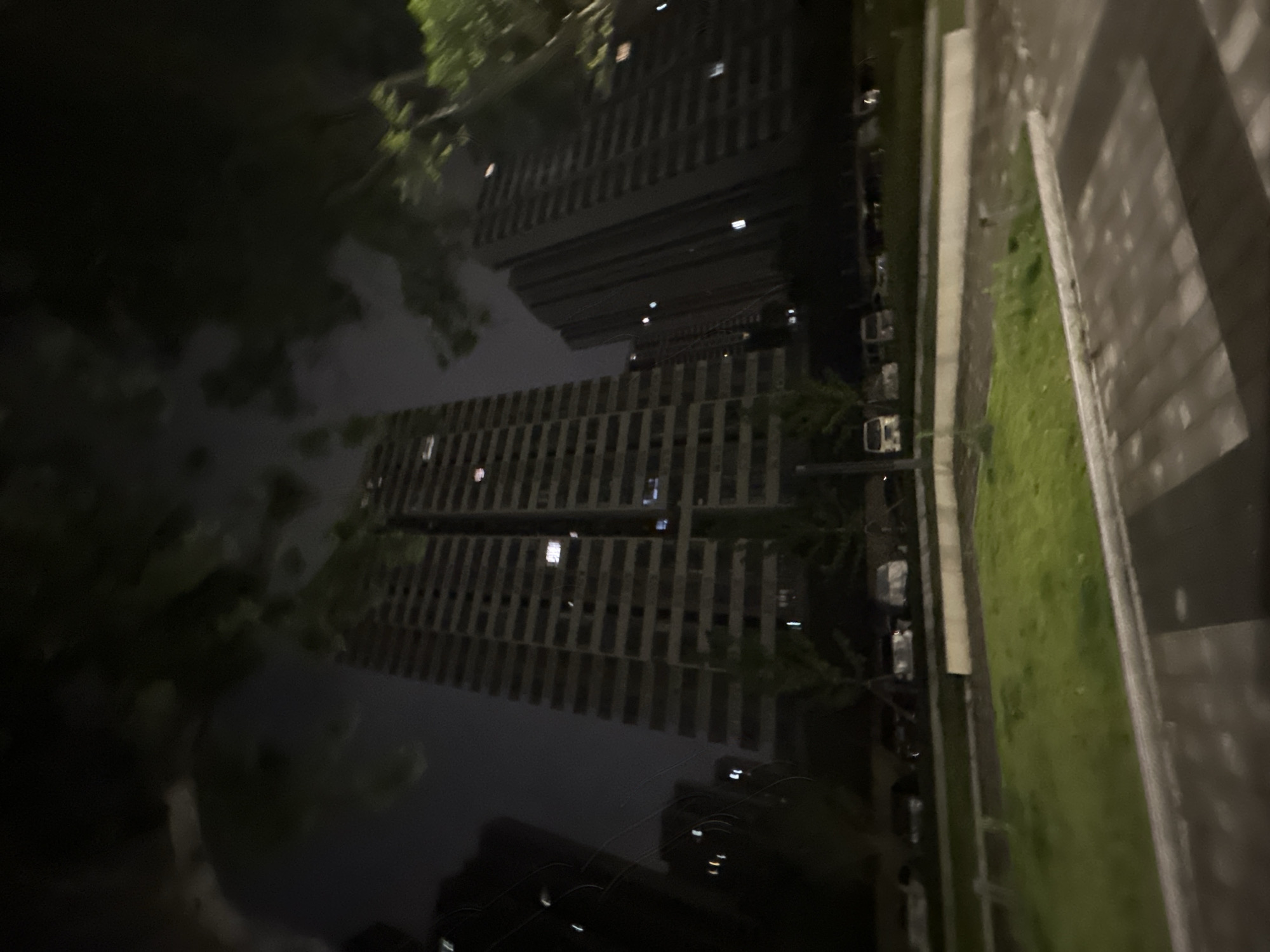 In the city of Chongqing High-tech Zone, China, at night, a view of two tall, rectangular apartment buildings dominates the scene. The buildings are constructed with a repetitive pattern of horizontal and vertical lines, creating a grid-like facade. Numerous windows are visible, some illuminated from within, suggesting occupancy. In the foreground, a patch of green grass is illuminated by soft light, bordered by a concrete walkway. Several cars are parked in the background, their forms partially obscured by shadows and the low light conditions. Trees with dark foliage are scattered throughout the scene, framing the buildings and adding depth to the composition. The overall atmosphere is dark and quiet, with the artificial lights of the buildings and streetlights casting a subdued glow on the urban landscape.