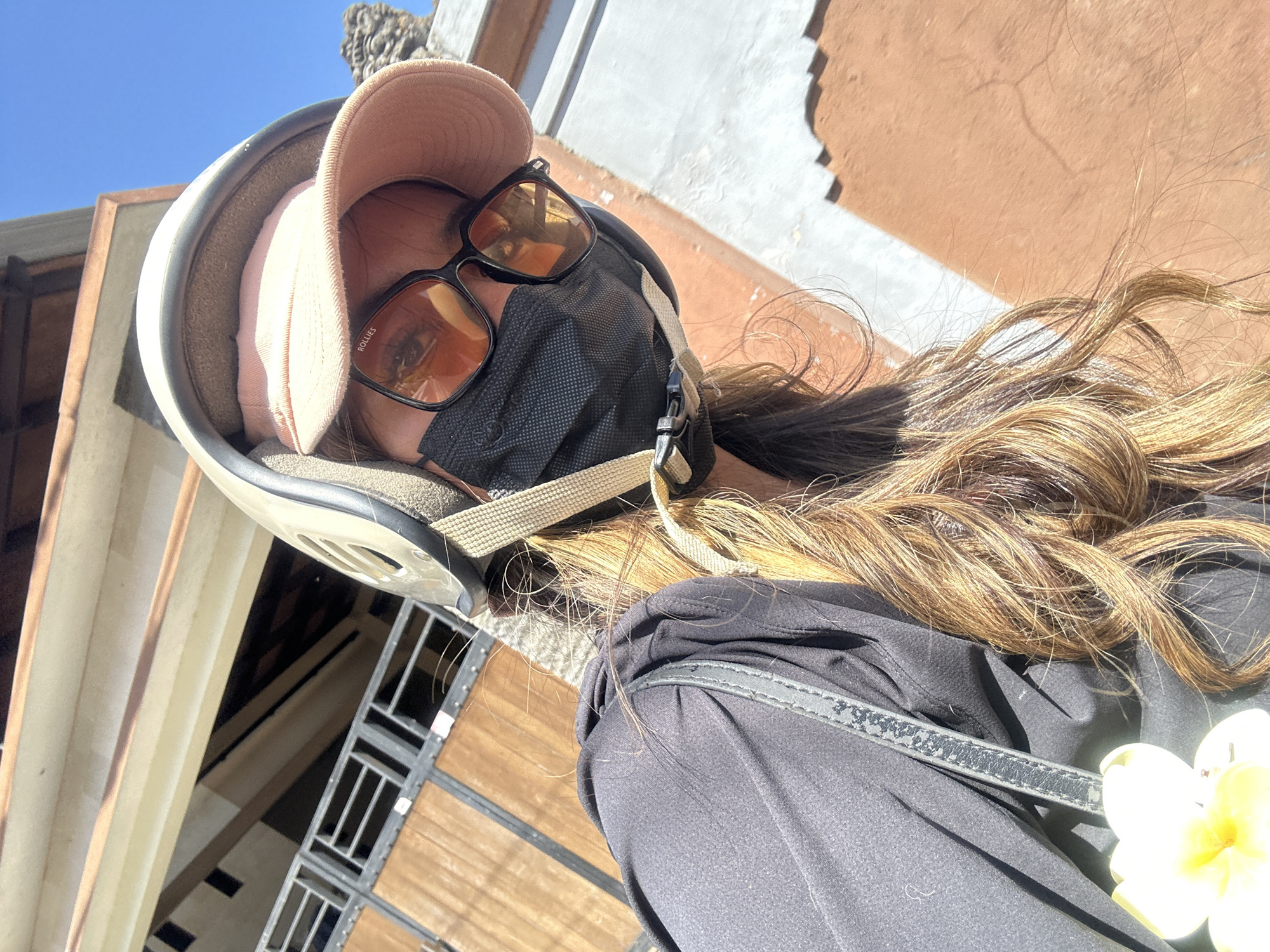 A selfie taken from a slightly high angle shows a woman wearing a tan helmet, a light pink baseball cap, dark sunglasses, and a black face mask. Her long, wavy brown hair is visible. She is wearing a black top and a white flower is pinned to her clothing on the left side of the frame. The background includes a building with wooden elements and a clear blue sky. The setting appears to be outdoors, possibly on a street in Denpasar, Indonesia, suggested by the architectural style and tropical flora. The lighting indicates it is daytime.