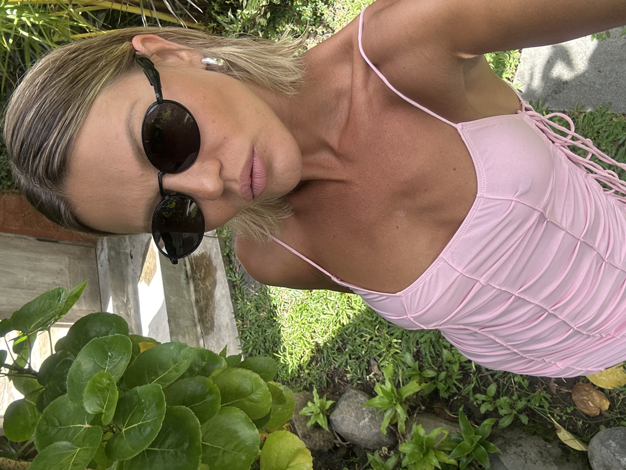 A woman wearing sunglasses and a pink dress poses outdoors. The environment is a garden with greenery. The location is in North Kuta, Indonesia.