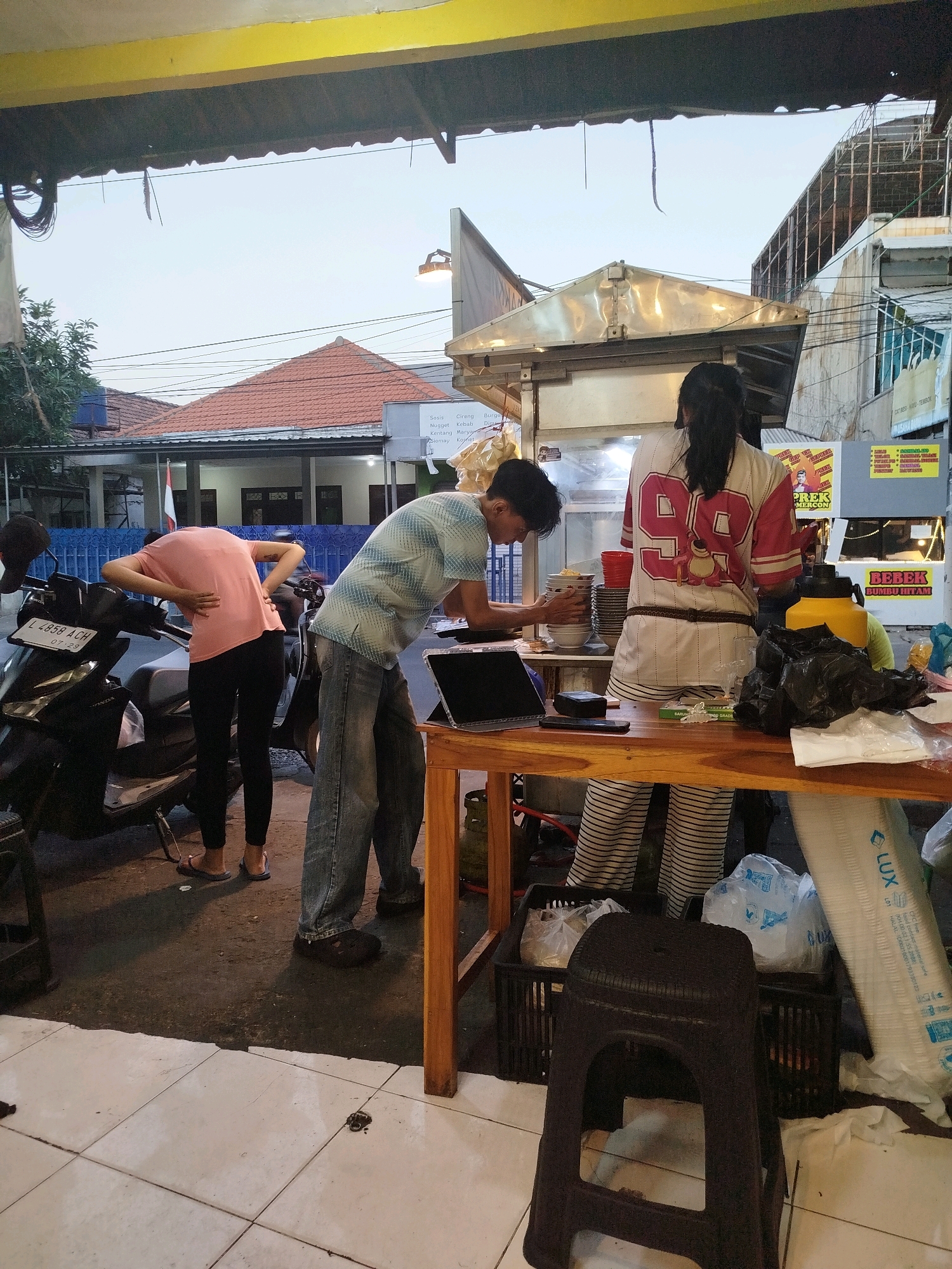 The image depicts an outdoor food stall scene in Surabaya, Indonesia, likely in the evening. Three individuals are present. A person wearing a pink t-shirt and black pants is bent over, facing away from the camera, with their hands on their lower back, suggesting discomfort or exertion. They are standing next to a black scooter with a license plate reading "L 2858 ACH". Another person, a male, is standing behind a food counter, wearing a blue and white striped shirt and jeans, and appears to be preparing food or organizing dishes. He is looking down at a laptop and bowls of food. A third individual, a female, is standing behind the counter, facing away from the camera, wearing a white shirt with red trim and the number "99" on the back, along with striped pants. The food stall has a metal roof and appears to be set up along a street. Various food items, bags, and cooking equipment are visible on and around the counter. The background shows buildings and utility wires, typical of an urban environment. The lighting suggests it is either early morning or late evening.