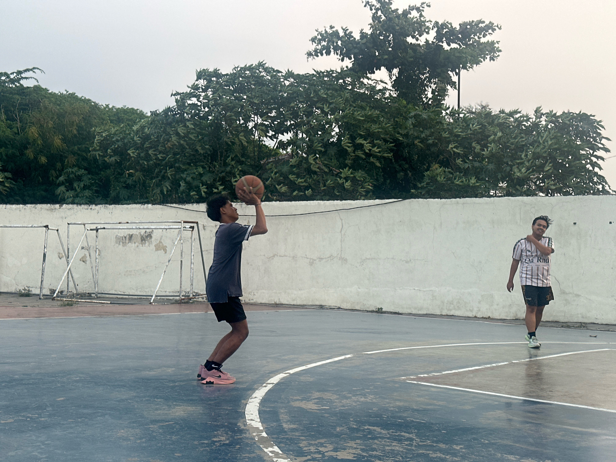 The image depicts two young men on an outdoor basketball court. The court surface is dark blue with white lines. In the background, a white concrete wall runs horizontally, topped by lush green trees against an overcast sky. To the left, partially visible behind the wall, are white metal structures resembling goal frames, possibly from a volleyball or badminton court.
The man on the left is in mid-action, preparing to shoot a basketball. He is wearing a dark grey t-shirt and black shorts, with pink and black athletic shoes. His body is angled, with his right arm raised, holding the basketball, and his left arm extended for balance. He appears to be of East Asian descent with short, dark hair.
The man on the right is standing further down the court, facing towards the viewer and slightly to his left. He is wearing a striped t-shirt with black and white stripes and black shorts, with white and black athletic shoes. He is smiling and has his right arm bent at the elbow, with his hand near his face. He also appears to be of East Asian descent with short, dark hair.
The lighting suggests it is daytime, possibly late afternoon, with diffused light due to the cloudy conditions. The overall scene captures a casual moment of recreational activity on a basketball court. The specific location in Mojokerto, Indonesia, is not visually identifiable within the frame itself, but the description of the environment and people is consistent with common urban or suburban settings in the region.