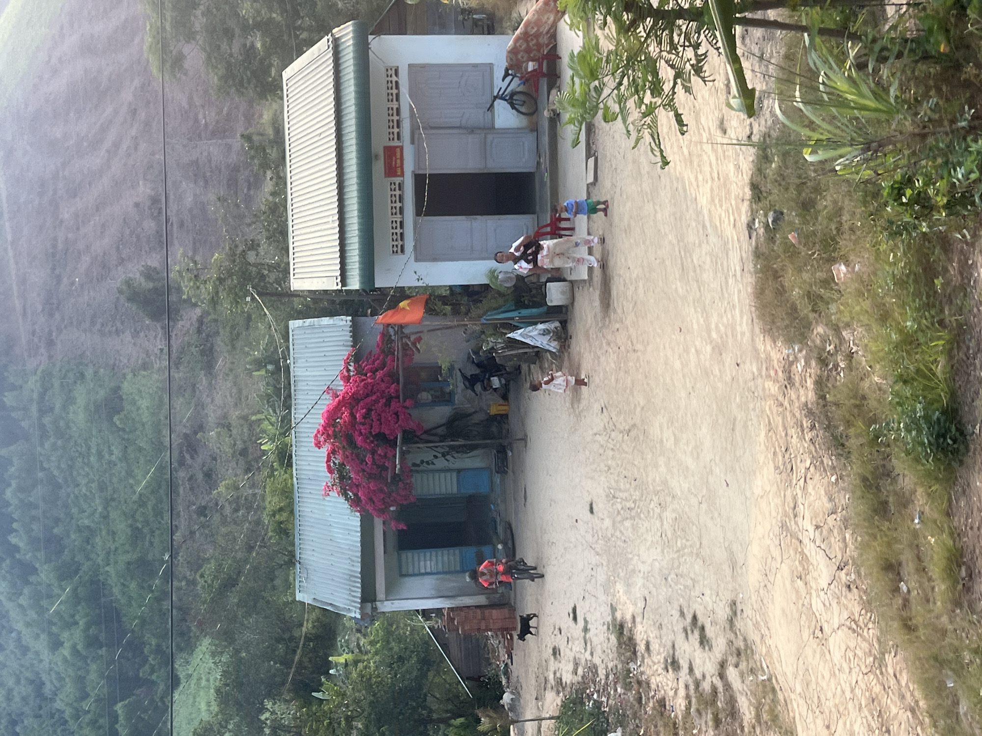 The scene depicts two single-story buildings in what appears to be a rural area of Myui, Vietnam. The building to the left is partially obscured by a large, flowering bougainvillea bush. The building to the right has an open doorway, and two adults and two children are standing in front of it. A woman on a bicycle, wearing an orange shirt, is riding on a dirt path in front of the other building. There is a dog running beside the bicycle. A motorcycle is parked in front of the building with the bougainvillea bush. In the background, there is a steep, green hillside covered in trees. The overall environment seems warm and sunny. Power lines run across the image.