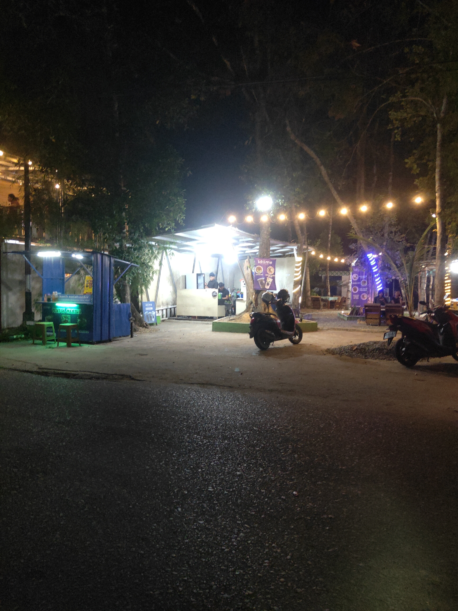 The image depicts a street scene at night in Sintang, Indonesia. A small food stall, with a blue facade and a green awning, is visible on the left. It displays the text "KAMI SUDAH ORDER" and "PENTOL MERTUA". Two green stools are placed in front of the stall. Further into the background, a larger, open-sided structure with a white roof is illuminated by warm string lights and a blue LED strip. Several people are present, some standing and some seated. Two motorcycles are parked in the foreground, one black and the other red. The ground is paved and appears to be wet in places. Trees with visible branches and leaves are scattered throughout the scene, suggesting an outdoor setting.