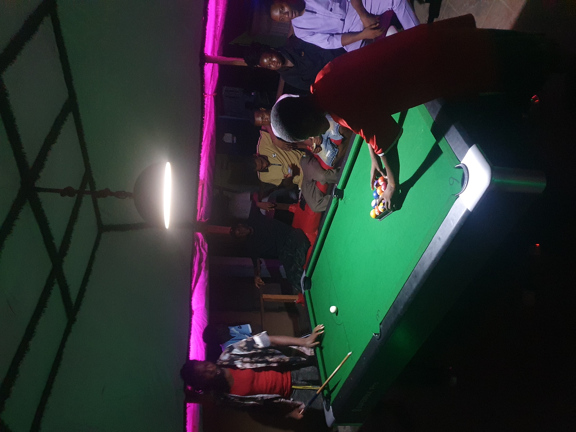 The image captures a nighttime scene inside a recreation area in Lokoja, Nigeria, where several individuals are gathered around a pool table. The setting is characterized by artificial lighting, including a bright vertical light fixture on the left and a vibrant pink neon light strip along a wall or ceiling edge, casting a distinct hue.
In the foreground, a person wearing a red t-shirt is actively arranging colorful billiard balls in a rack on a green-felted pool table, indicating the preparation for a game. A white cue ball and a cue stick are also visible on the table.
Multiple spectators are present around the table and in the background. To the left of the person racking balls, another individual in a striped shirt observes the table. Further back, several people are seated or standing, including one in a light purple, long-sleeved garment and another in a tan jacket featuring an "HH" logo. Others are partially visible in the dim lighting. The group appears engaged and relaxed, suggesting a social gathering centered around the game.
The ceiling above features a geometric structural pattern. Shadows are prominent, adding depth to the scene. The overall atmosphere is casual and convivial. Faint text, including "OFFICIAL PRO" and the number "6," is subtly visible on the white trim of the pool table.