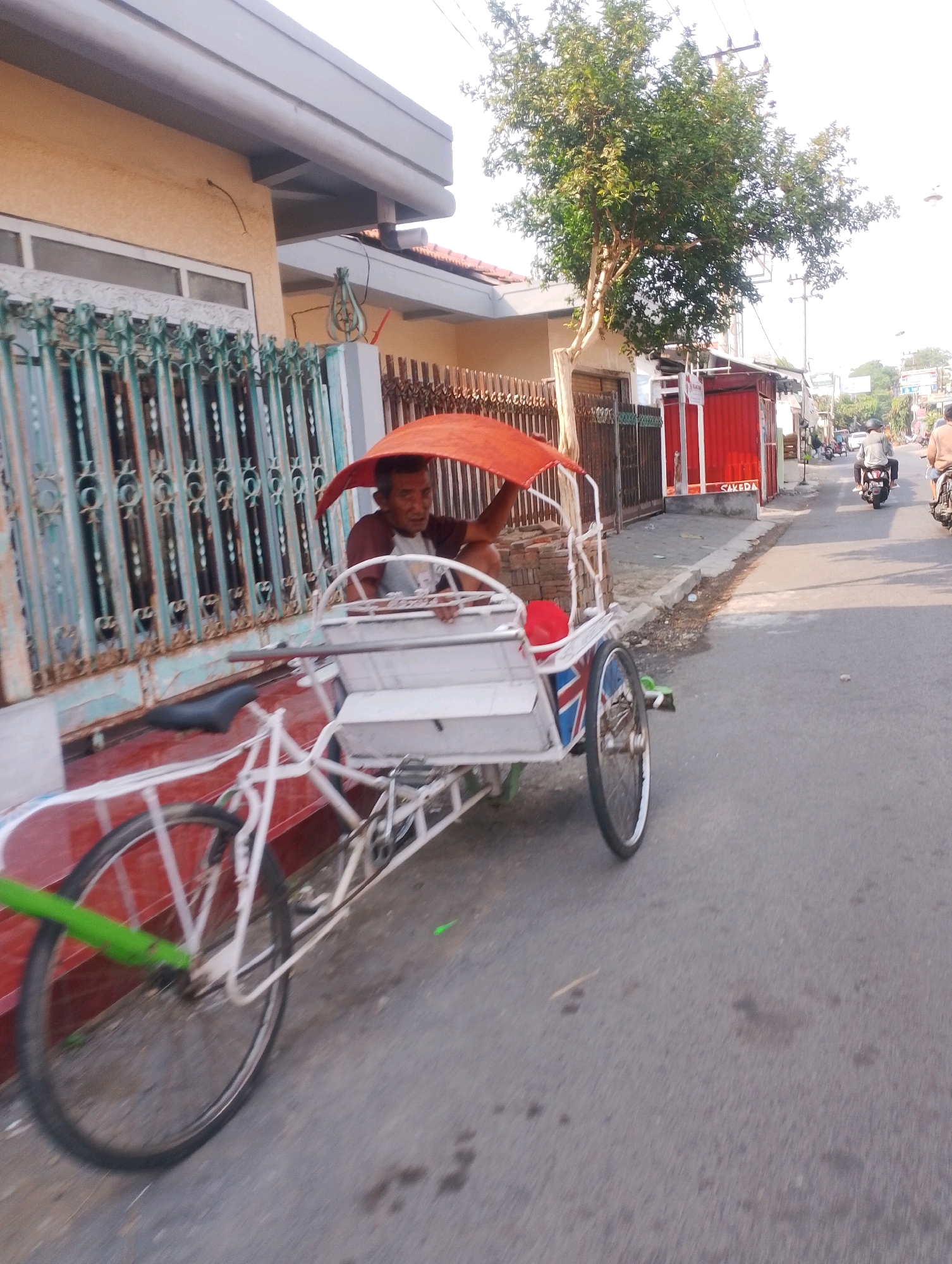 A middle-aged Indonesian man is sitting on a white and blue pedicab that is parked on the side of a paved road in Probolinggo, Indonesia. The man is wearing a maroon and grey shirt and appears to be looking at the camera. The pedicab has an orange canopy for shade and a front seat for passengers. The background shows a street with a few buildings, trees, and other vehicles, including motorcycles. The scene appears to be in a residential or commercial area, with houses and shops lining the street. The lighting suggests it is daytime, possibly late afternoon.