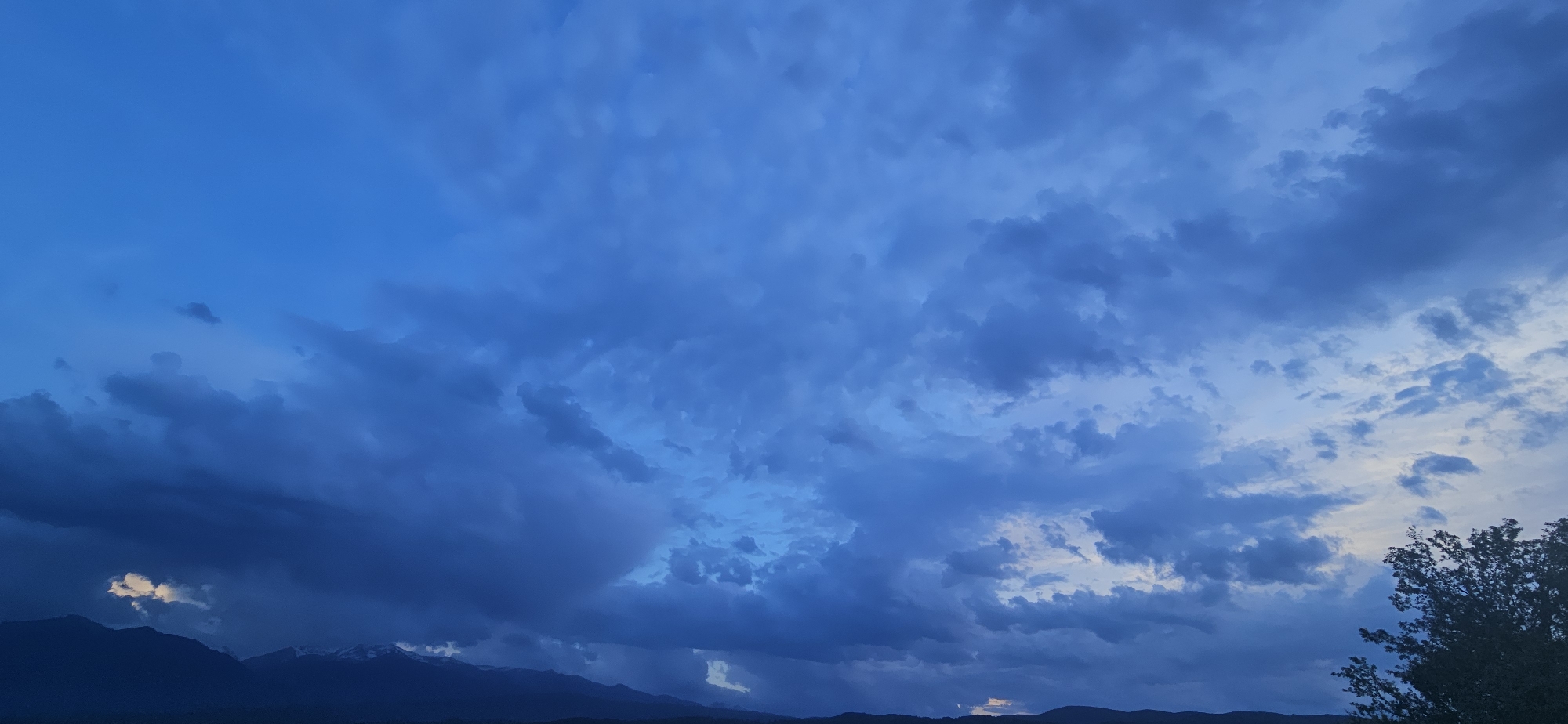 This media file captures a dramatic skyscape over a distant mountain range, likely during twilight hours, either dawn or dusk, given the low light and deep blue color palette. The entire sky is heavily overcast with a complex interplay of clouds.
The clouds themselves are varied, ranging from softer, more diffused layers in the upper and central parts of the sky to denser, darker, and more formidable cumuliform masses, especially in the lower-left and central regions. These dark clouds suggest significant atmospheric activity. Patches of lighter blue sky, some with hints of white, break through the cloud cover in various areas, notably in the upper-left, central-right, and lower-right, offering contrast to the prevailing dark tones.
Across the horizon, a dark, silhouetted mountain range is visible. Faint traces of snow or ice caps can be discerned on some of the higher peaks, particularly in the lower-left portion of the main image. A small, distinct patch of bright light or an illuminated cloud appears low in the sky, just above the mountains on the left side. On the far right edge, the dark foliage of a tree canopy is partially visible.
The scene emphasizes natural elements with no human subjects, specific activities, or visible text. The overall impression is one of natural grandeur and dynamic weather. The location context provided is Gutsal, Bulgaria.