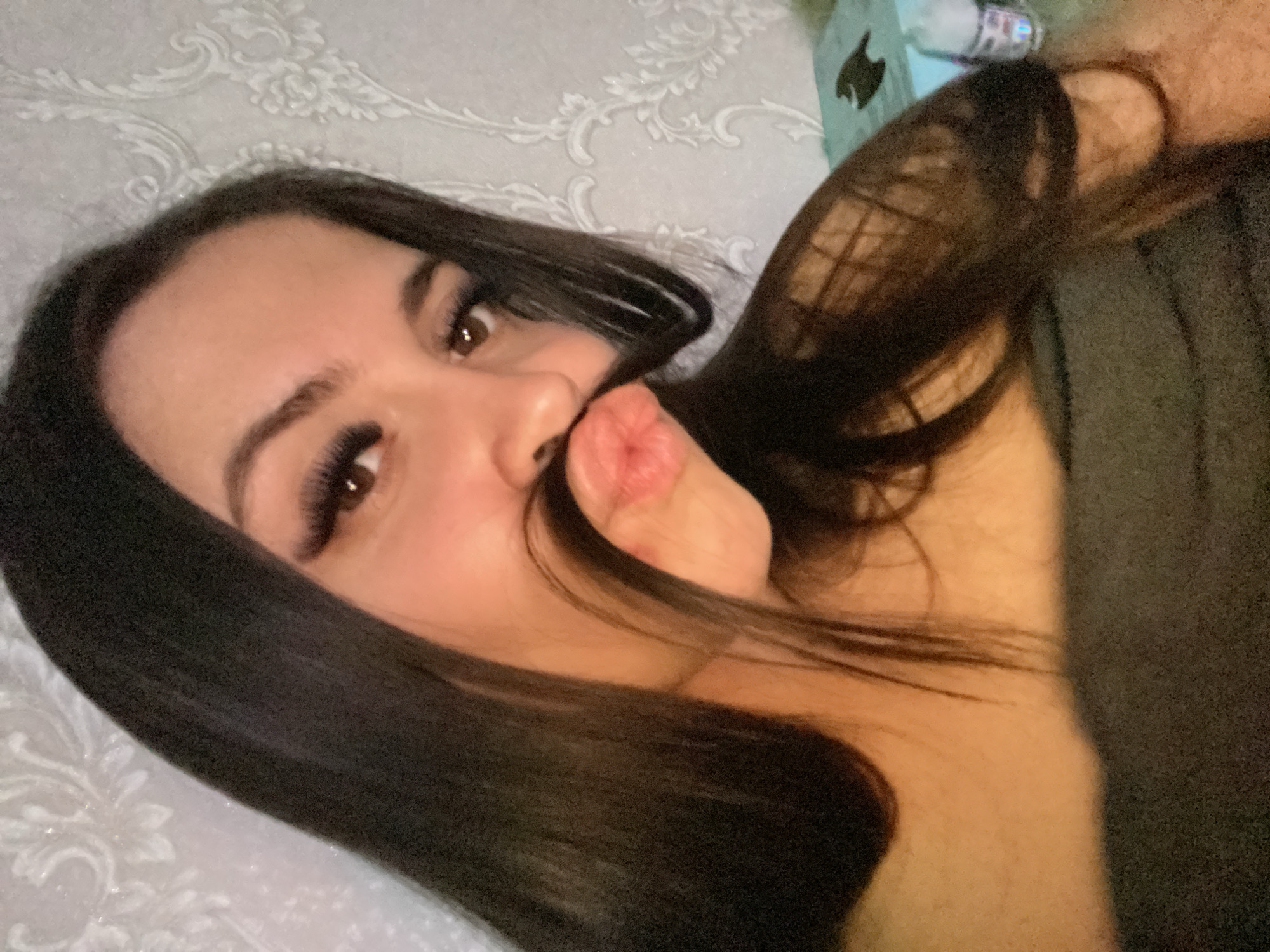 A young woman with long, dark hair is playfully posing for a selfie while lying on a white textured surface. She is holding strands of her hair to resemble a mustache above her puckered lips. Her eyes are brown with long lashes. A box with a black silhouette, likely the Apple logo, and a water bottle are visible in the background. The location, Chaikovsky, Russia, cannot be verified from the image.