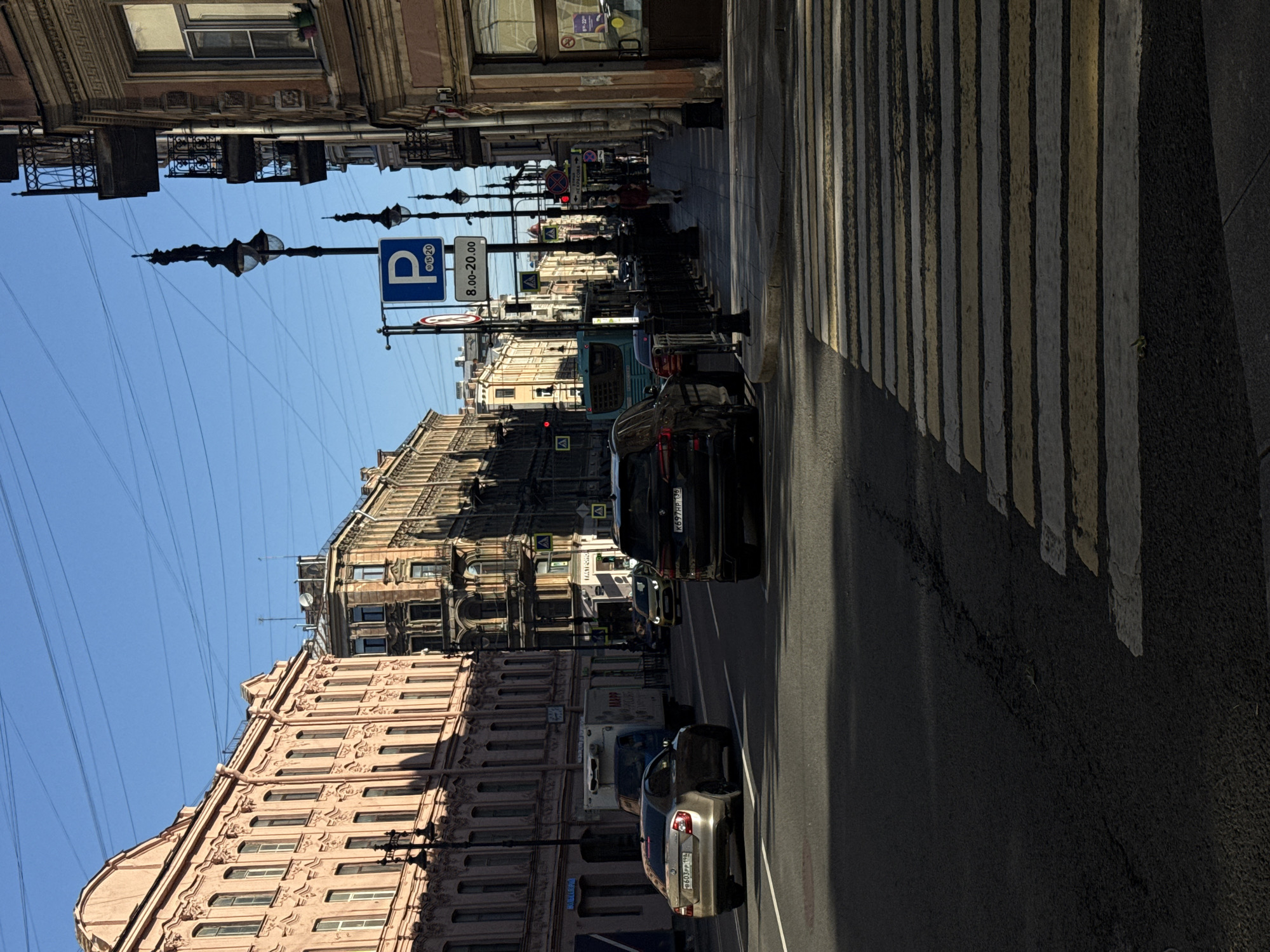 The image depicts a street scene in Saint Petersburg, Russia, with the distinctive architecture of the city visible on both sides of the road. On the left, a light pink building with ornate detailing lines the street. Across from it, on the right, a building with a darker facade and balconies is visible. The street is paved and shows a pedestrian crossing with yellow and white stripes. Several vehicles are present, including a gold sedan, a dark SUV, and a white truck. A blue bus is also in motion. A parking sign with the letter 'P' and time restrictions (8.00-20.00) is mounted on a lamppost. Overhead wires crisscross the clear blue sky. The lighting suggests it is daytime, with shadows cast across the road and sidewalk. A lone pedestrian is walking on the sidewalk on the right side of the image.