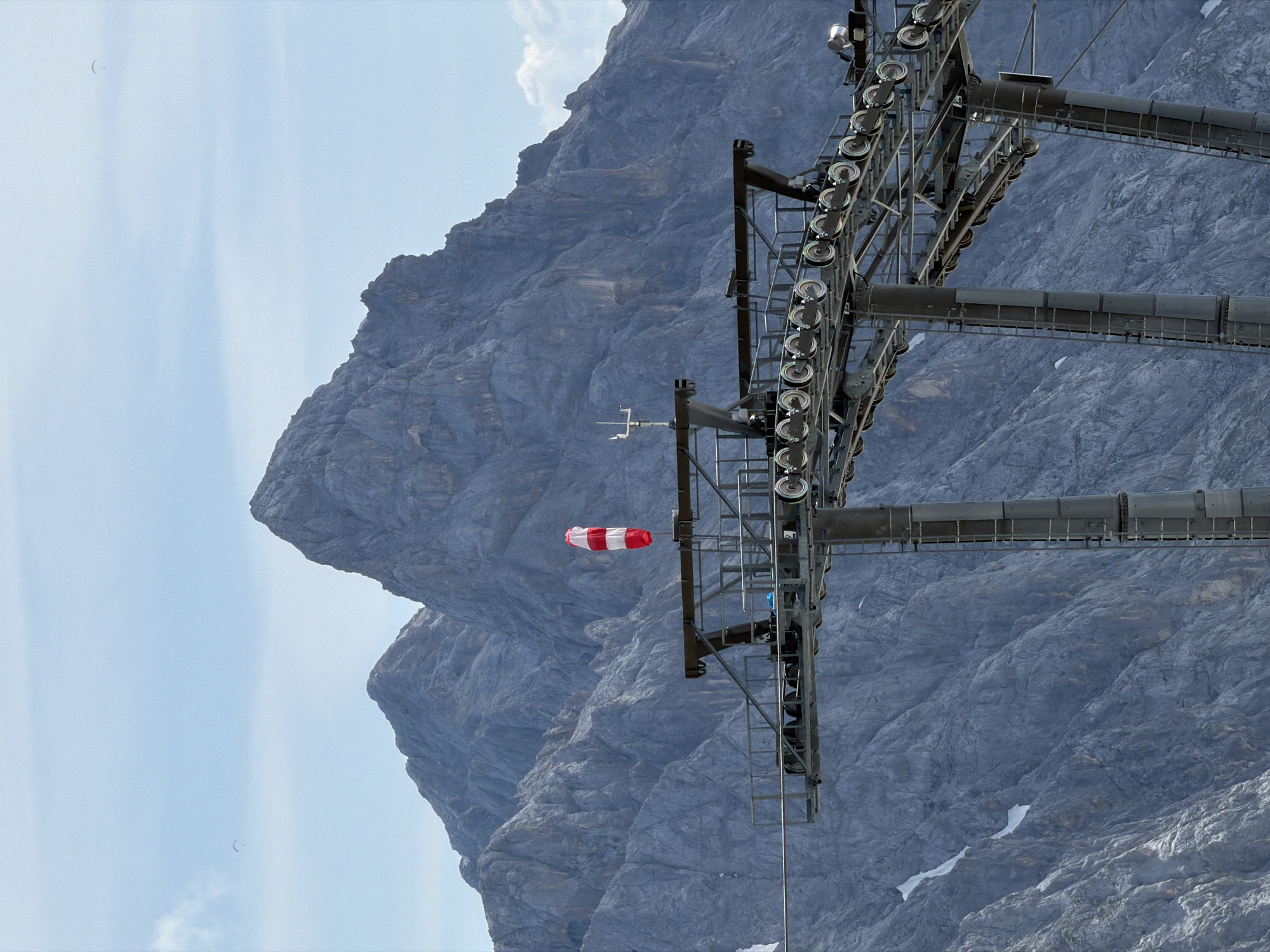 In Garmisch-Partenkirchen, Germany, a ski lift tower stands prominently against a backdrop of majestic, rugged mountains. The tower supports a complex network of cables and pulleys, indicating its role in the ski resort's operations. A windsock, striped in red and white, flutters atop the structure, suggesting windy conditions. In the distance, two paragliders can be seen soaring in the clear blue sky, adding a sense of adventure to the serene mountain landscape. The scene captures the essence of a high-altitude alpine environment, where human engineering intersects with the raw beauty of nature.