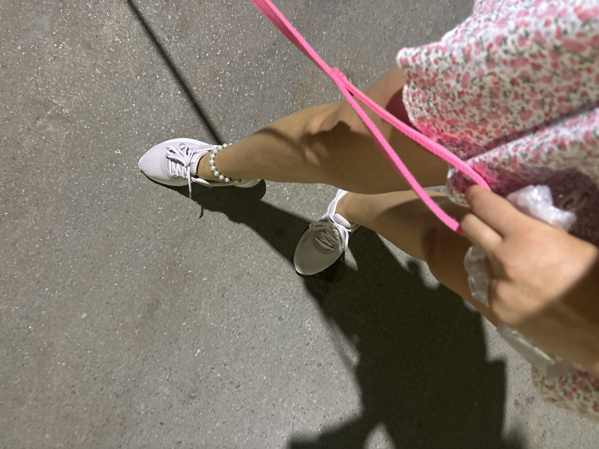 The image shows a person's legs and feet walking on a gray asphalt surface, likely a street or sidewalk, in Budva, Montenegro. The person is wearing light pink athletic shoes and a floral-patterned dress. A bright pink strap, possibly from a bag or accessory, is visible across the foreground. The lighting suggests it is either dusk or night, as a shadow is cast by the person, indicating a light source from the side. The ground texture is rough and uneven, typical of urban pavement.