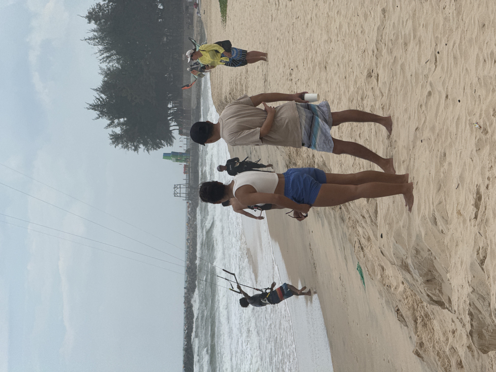The image depicts a beach in Mũi Né, Vietnam, with several individuals engaged in or observing kite surfing. In the foreground, two people stand on the sandy beach, facing away from the camera. The person on the left wears a white crop top and blue shorts, while the person on the right wears a light brown t-shirt and striped shorts, holding a white cup. To the left, closer to the water, a person in a dark shirt and striped shorts is holding a kiteboarding bar, appearing to be preparing for or engaged in kite surfing. Waves are breaking on the shore. Further down the beach, another person in a black wetsuit is visible, and a woman in a yellow top and patterned skirt is standing near kiteboarding equipment. In the background, trees and a structure, possibly a pier or observation tower, are visible. The sky is overcast.