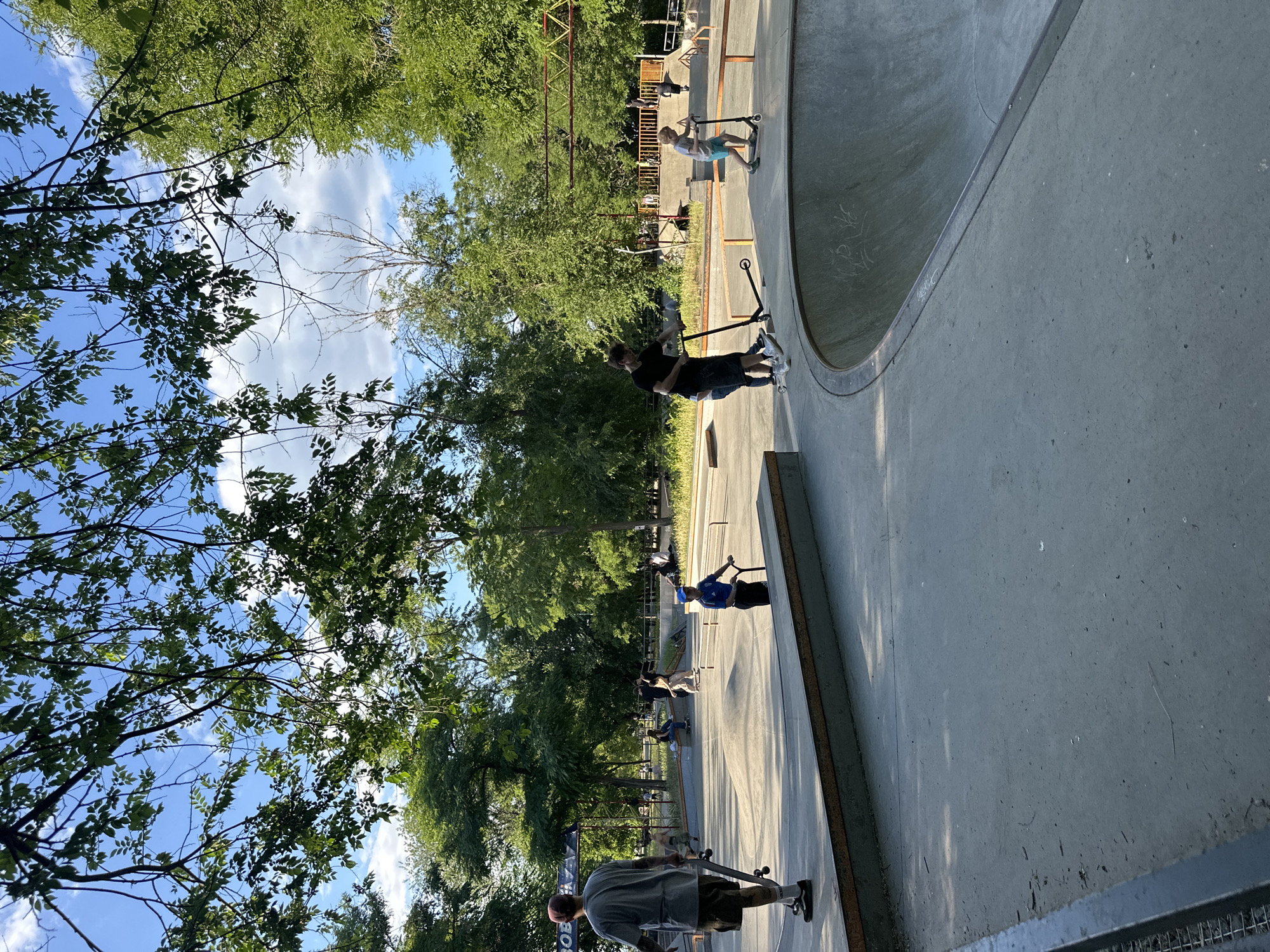 An outdoor concrete skatepark is pictured on a sunny day. Green trees surround the park, and the sky above is blue with scattered clouds. Several people are engaged in activities within the skatepark.
In the foreground, a person wearing a grey t-shirt and brown shorts walks away from the camera, holding a skateboard. To the right, a young person in a blue t-shirt and blue cap is riding a scooter. Further into the park, a person in a black t-shirt and shorts is also riding a scooter. Another young person in a grey t-shirt and blue shorts is on a scooter, riding towards the foreground.
In the background, there are ramps, ledges, and a bowl feature typical of a skatepark. Other individuals are visible sitting and standing around the park. A structure with a yellow and orange geometric design is visible in the middle ground. The ground is primarily concrete with some patches of dirt and debris.