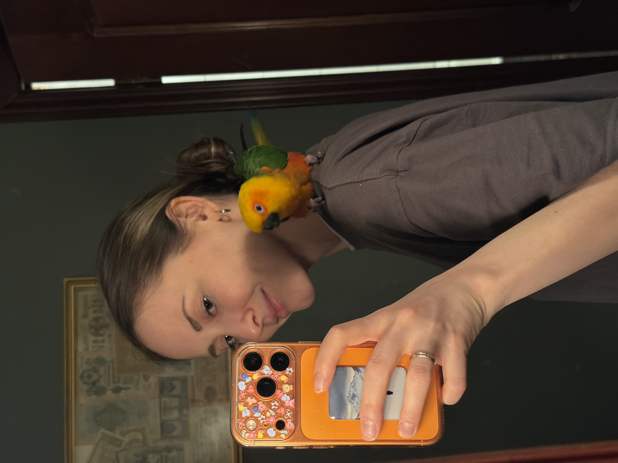A woman is taking a selfie in a mirror. A colorful parrot sits on her left shoulder. She holds an orange smartphone with a decorative case in her right hand. A picture frame is visible in the mirror behind her. The location cannot be determined from the image.