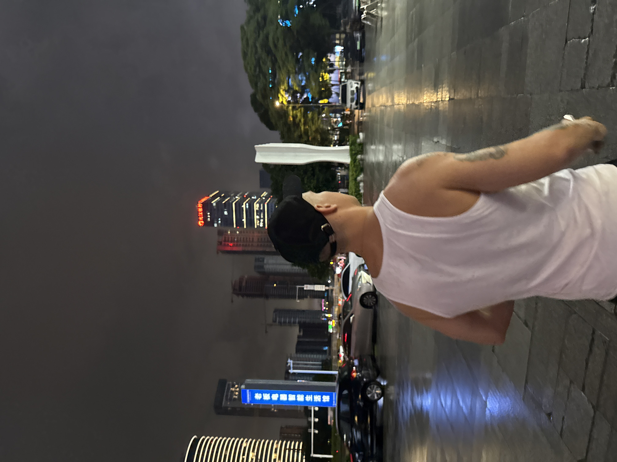A person wearing a white tank top and a black baseball cap is standing in an outdoor plaza at night. The plaza appears to be wet, with reflections from lights on the ground. In the background, there are several tall buildings, some illuminated with red and white lights. One building has Chinese characters on it. A white sculpture is also visible in the background, along with trees. Several cars are parked and driving in the scene, and streetlights are on. The image is likely taken in Tianhe District, China, given the presence of modern architecture and signage in Chinese.