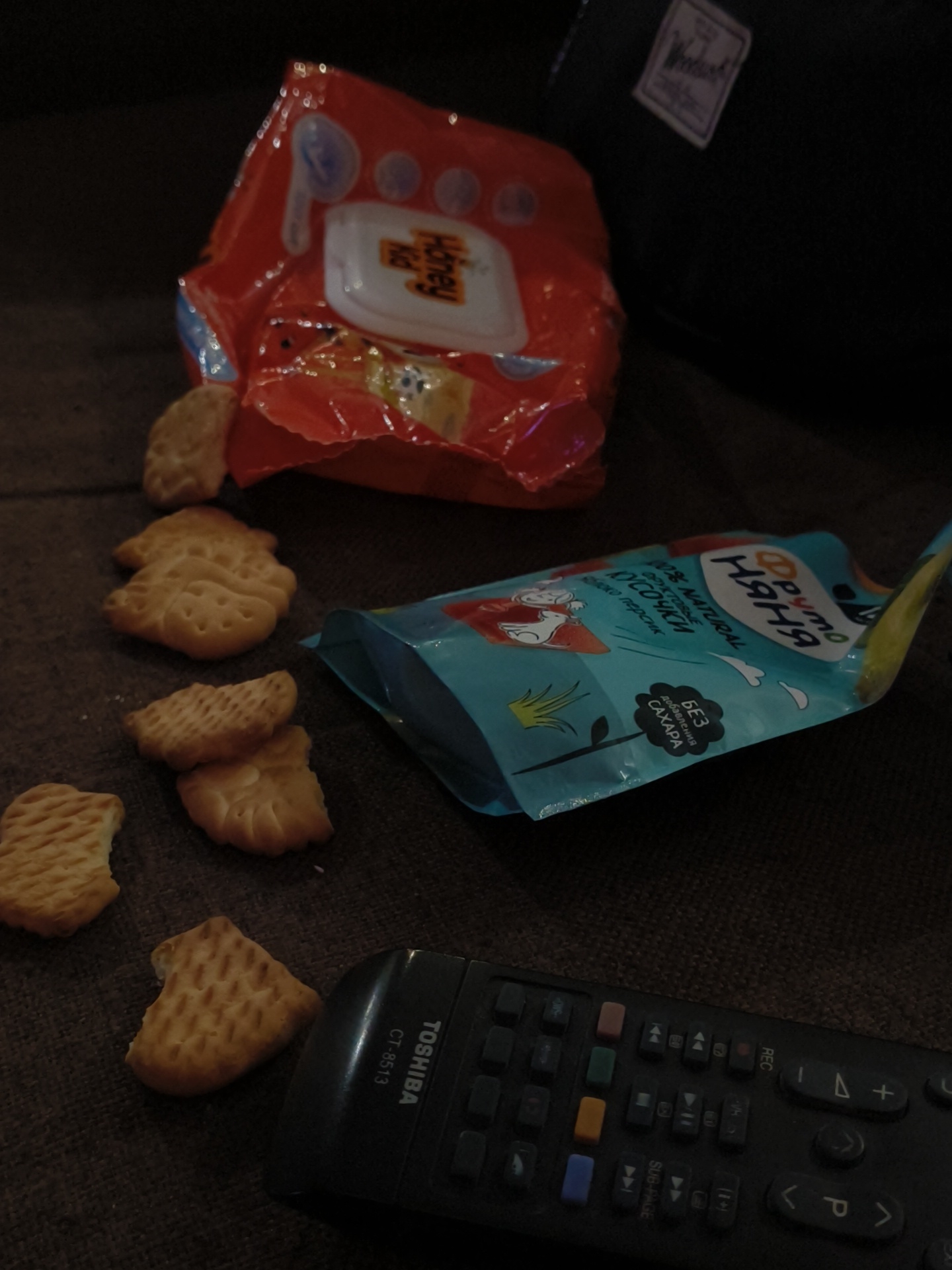 The image depicts a dimly lit indoor scene, likely in a living room in Chaikovsky, Russia. On a dark brown, textured surface, several items are scattered, suggesting a casual moment of relaxation and snacking.
In the foreground, a black Toshiba TV remote control, model CT-8513, is partially visible. Scattered across the surface are several light-colored, patterned crackers or biscuits; some are rectangular, while others have distinct shapes, including one that resembles an animal.
Near the center, a crumpled blue food pouch for children is present. Visible text on the pouch includes the Russian brand "Фруто Няня" (Fruto Nyanya), "ФРУКТОВЫЕ КУСОЧКИ" (Fruit Pieces), "яблоко персик" (apple peach), "100% NATURAL", and "БЕЗ добавления САХАРА" (NO added SUGAR).
Further back, a red, crumpled package of "Honey Kid" wet wipes with a clear opening flap is seen. One cracker is also near this package. In the upper right corner, a dark bag or backpack with a white rectangular label, possibly reading "Herschel", is partially visible. The dim lighting suggests the scene takes place in the evening or at night. The scattered snacks and remote control imply someone is unwinding, possibly while watching television.