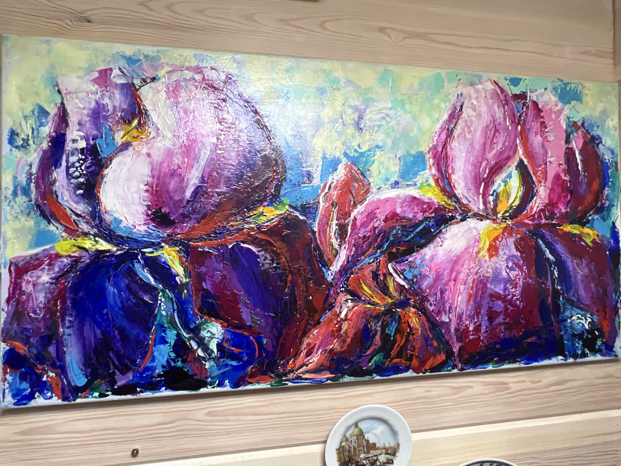 The image shows a close-up, side view of a horizontal, impasto oil painting of irises, displayed on a light-colored wooden wall. The painting is vibrant and abstract, with thick brushstrokes of pink, purple, blue, red, and yellow paint creating a textured depiction of the flowers. The background is a wash of light green and blue. Below the painting, on the wooden wall, are two small decorative plates. One is visible in the foreground and features an image of a European cityscape, possibly Venice, with buildings and canals under a blue sky. The other plate is partially visible to the right. The wooden wall appears to be paneling with visible grain and knots. There are no people visible in the image. The environment suggests an indoor setting, possibly a home or an art gallery. The location of Angarskie Khutora, Russia, is not directly discernible from the visual elements of the painting or the surrounding objects; this information is external to the image content.