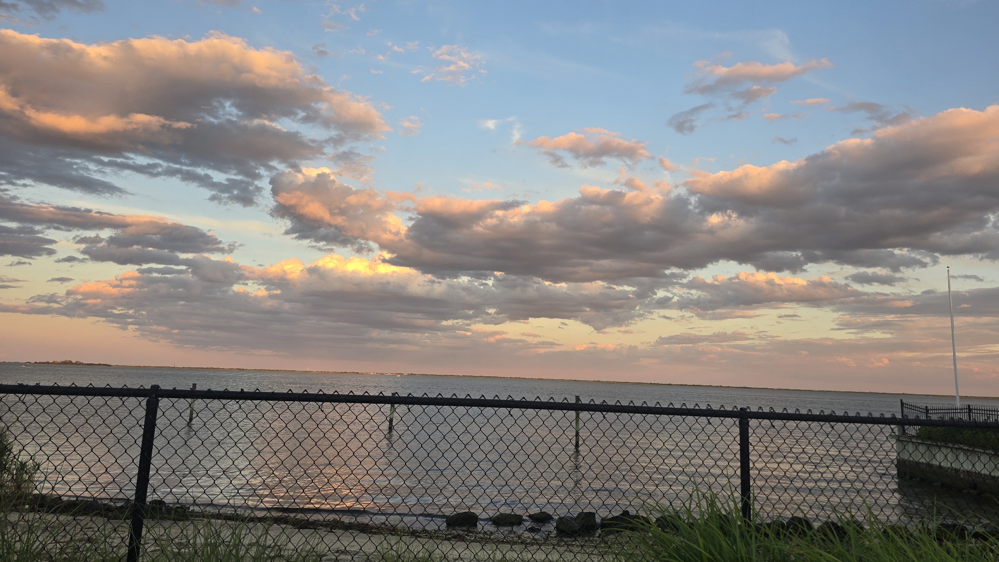 The image shows a sunset over the water in Copiague, New York. The sky is filled with clouds tinged with pink and orange from the setting sun, contrasting with the blue sky. A chain-link fence runs across the foreground, with tall green grass growing in front of it. Beyond the fence, a calm body of water stretches to the horizon, where a distant landmass is visible. Reflections of the colorful sky can be seen on the water's surface. A tall, thin flagpole stands to the right of the frame, with a small section of a paved walkway and railing visible behind the fence on the right side of the image.
