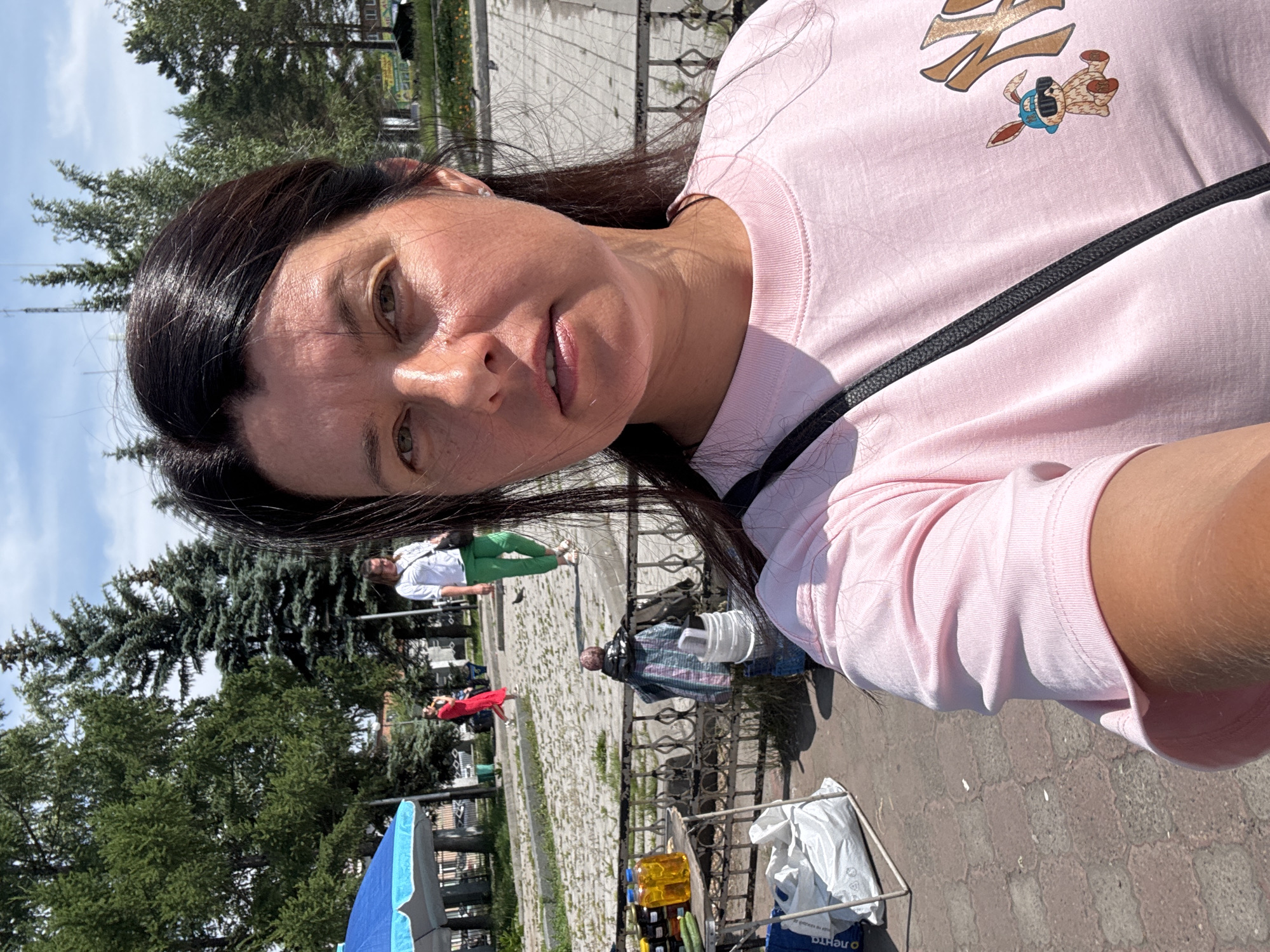 The image shows a woman taking a selfie in an outdoor public space, possibly a market or park, in Prokopyevsk, Russia. The woman is wearing a pink t-shirt with a graphic of a rabbit wearing sunglasses. Behind her, several other people are visible, including a woman in a white shirt and green pants and another in a red dress. There are trees, a blue umbrella, and a small table with bottles and vegetables. The ground is paved with interlocking stones. The sky is partly cloudy. The overall impression is of a bright, sunny day in a semi-urban environment.