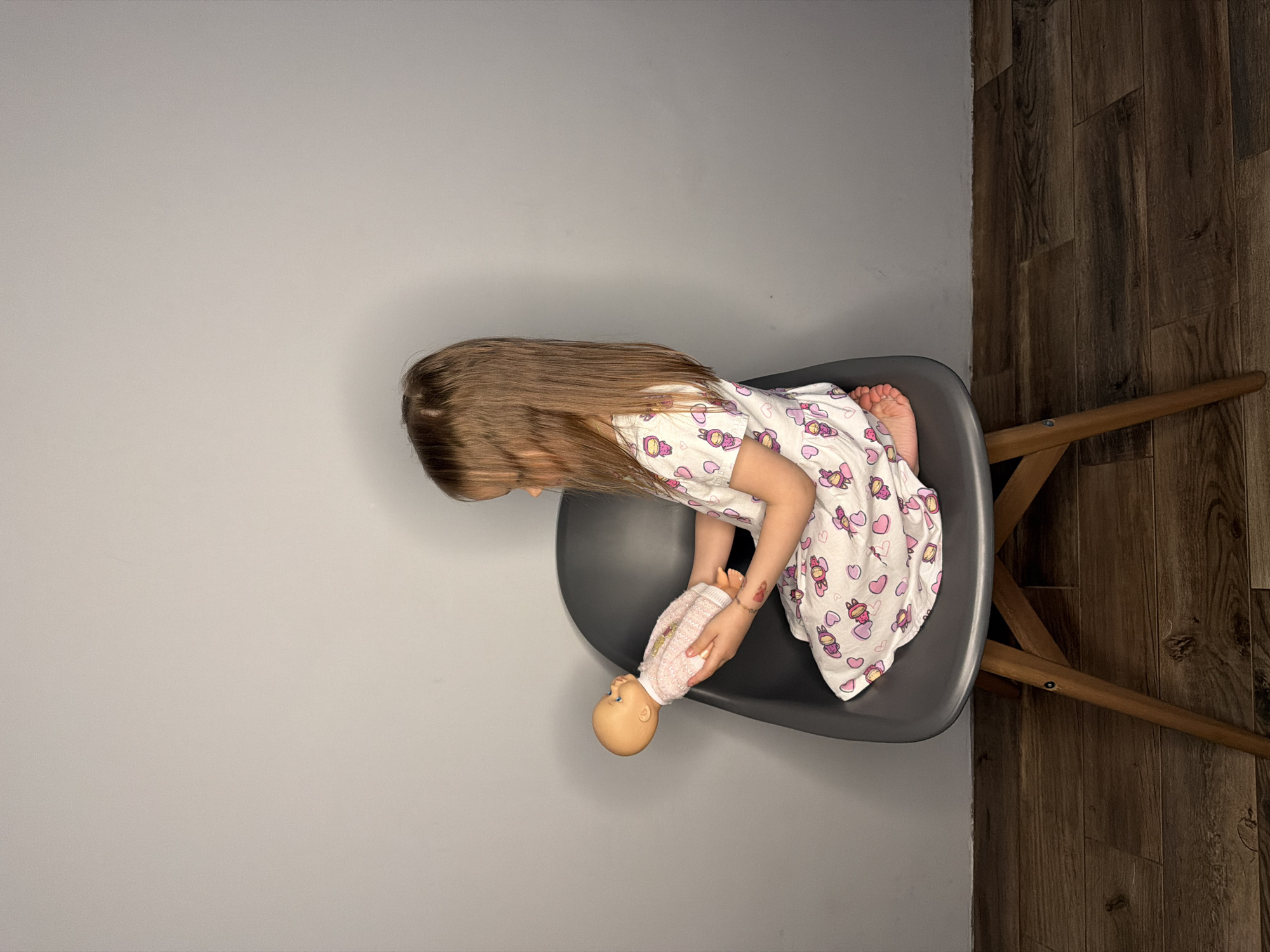 A young girl with long brown hair is sitting on a gray chair, holding a baby doll. She is wearing a white nightgown with cartoon images of girls and hearts. The chair is positioned at the intersection of a light gray wall and a dark wood-patterned floor. The setting appears to be indoors. The image was taken in Chaikovsky, Russia.